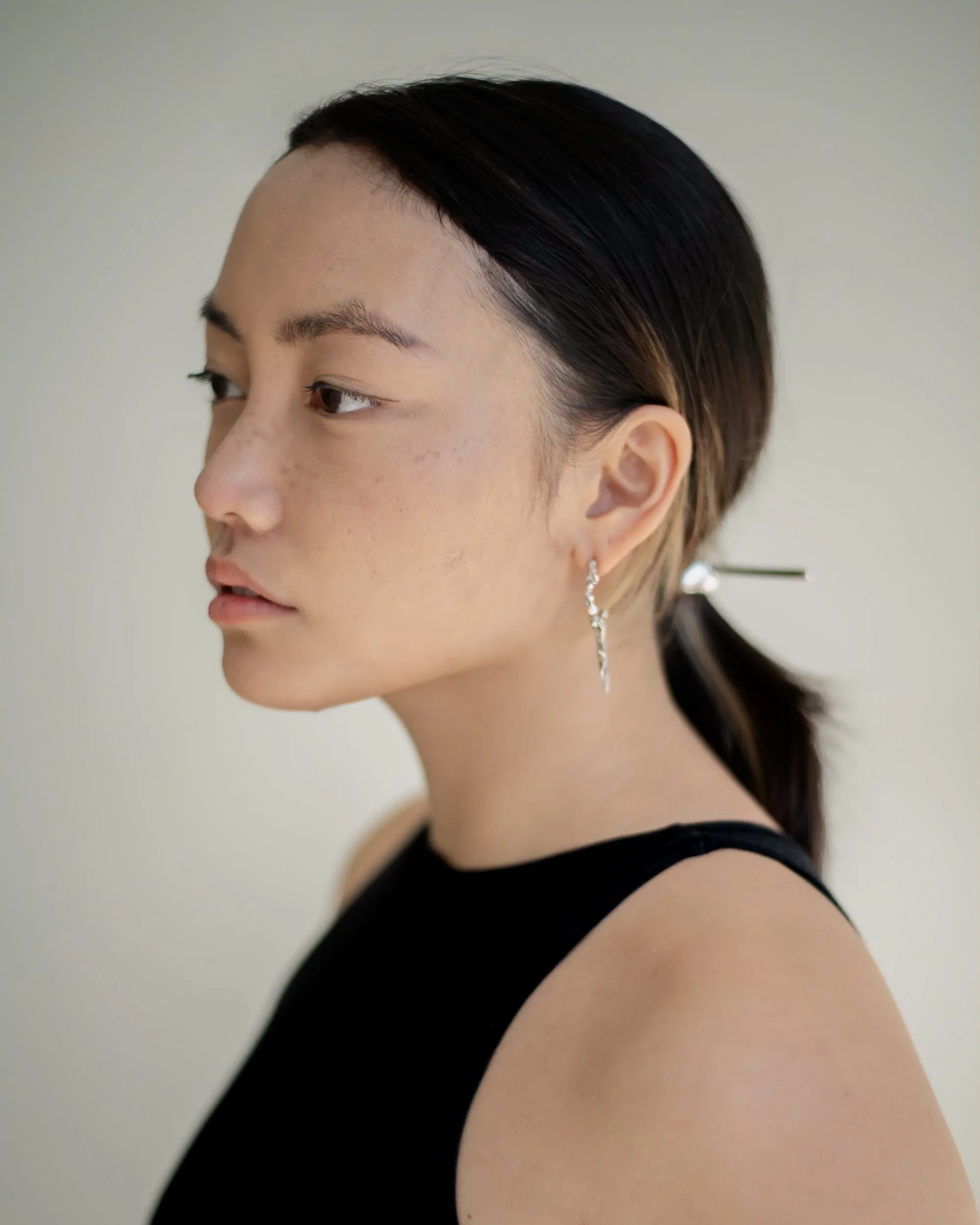 A close-up side profile of a young woman with dark hair tied back, wearing a black sleeveless top and a dangling earring, photographed during a portrait photography session in Los Angeles with fashion photographer Gí Tenamá.