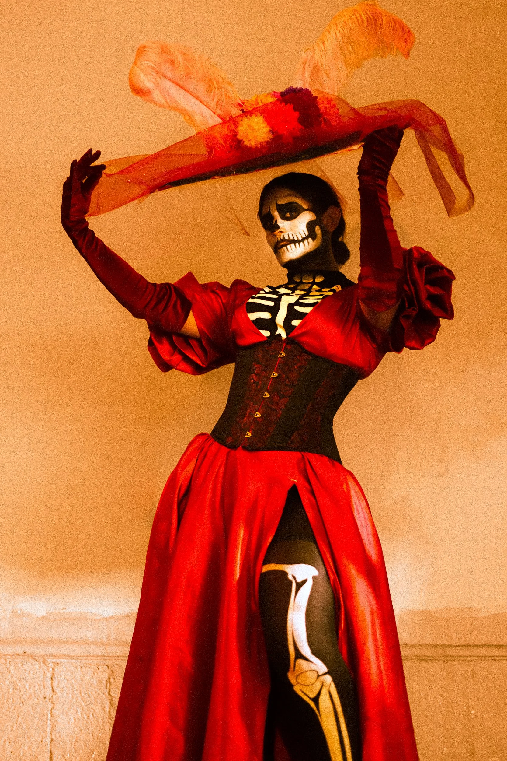 A person dressed in a Day of the Dead-inspired skeleton costume featuring a red dress with puffed sleeves, a black corset, and skeleton face paint, holding a large orange hat decorated with feathers and flowers.