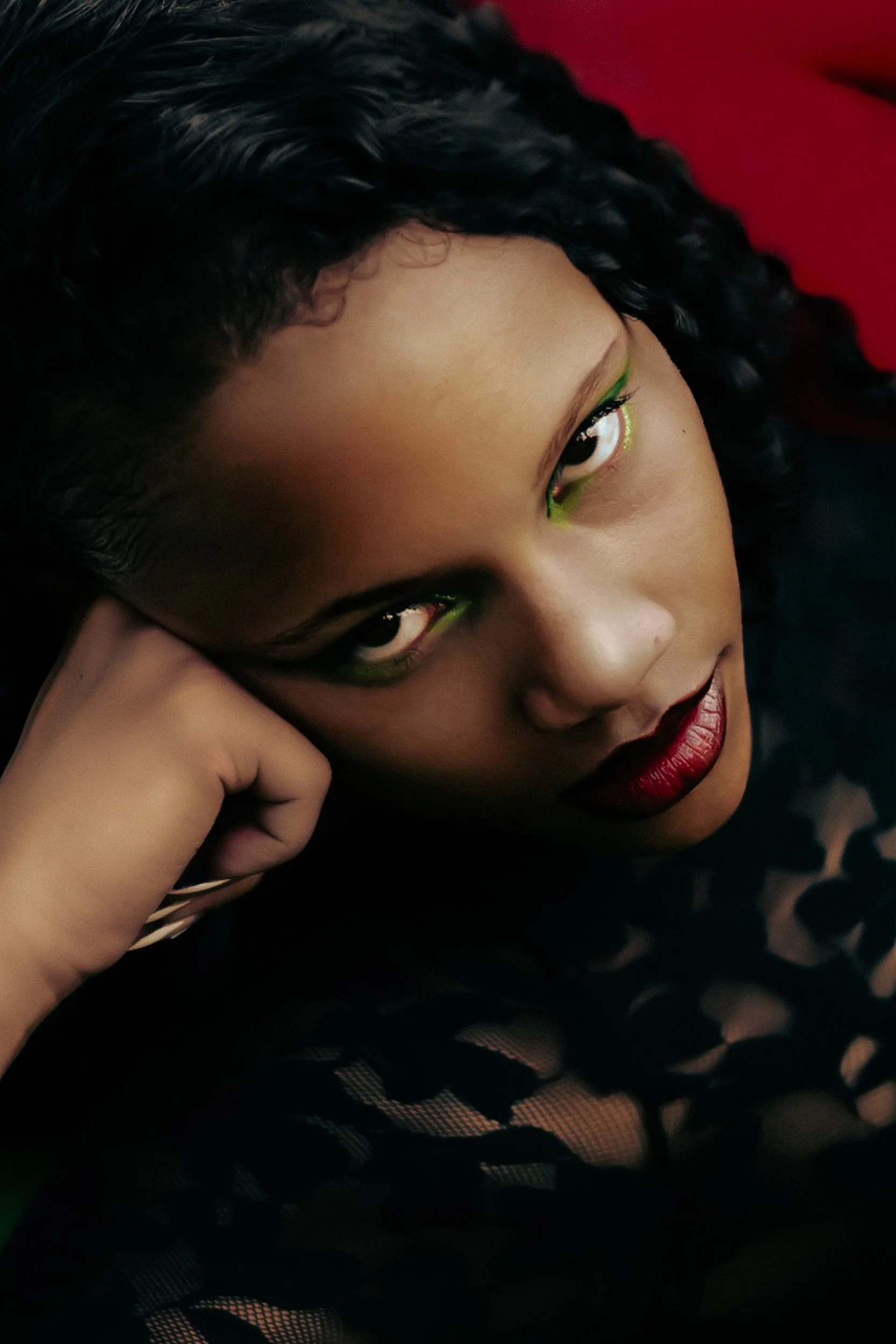 Close-up of a Black woman model with dark curly hair and bold makeup, resting her head on her hand, featuring lime green eye makeup and dark red lipstick, shot for an editorial fashion photography session in Los Angeles with fashion photographer Gí T