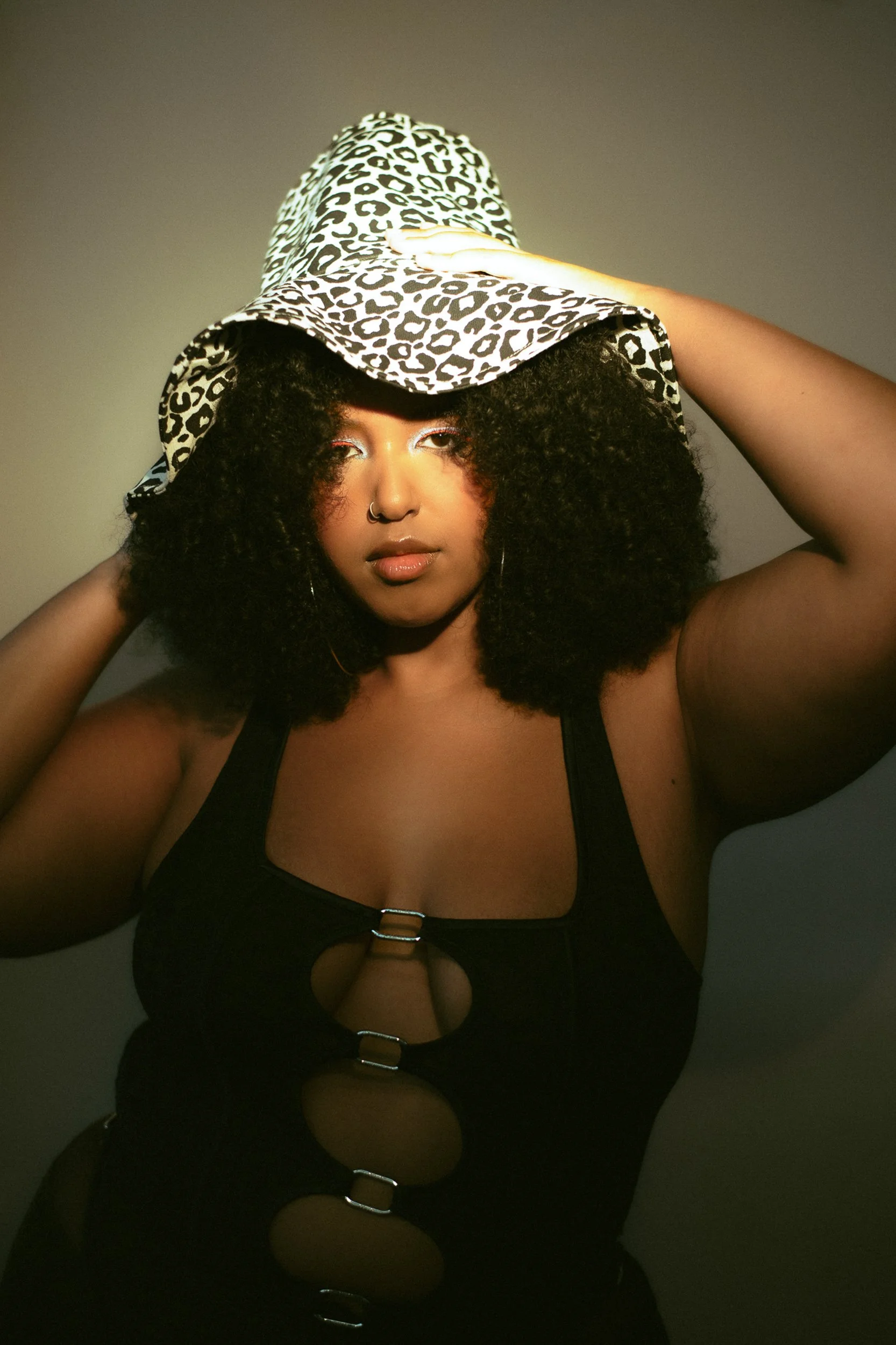 A woman with curly hair wearing a leopard print wide-brim hat, black top with large circular cutouts, and hoop earrings. She touches her hat with one hand and looks into the camera, A woman with curly black hair wearing bright makeup, earrings, a rai