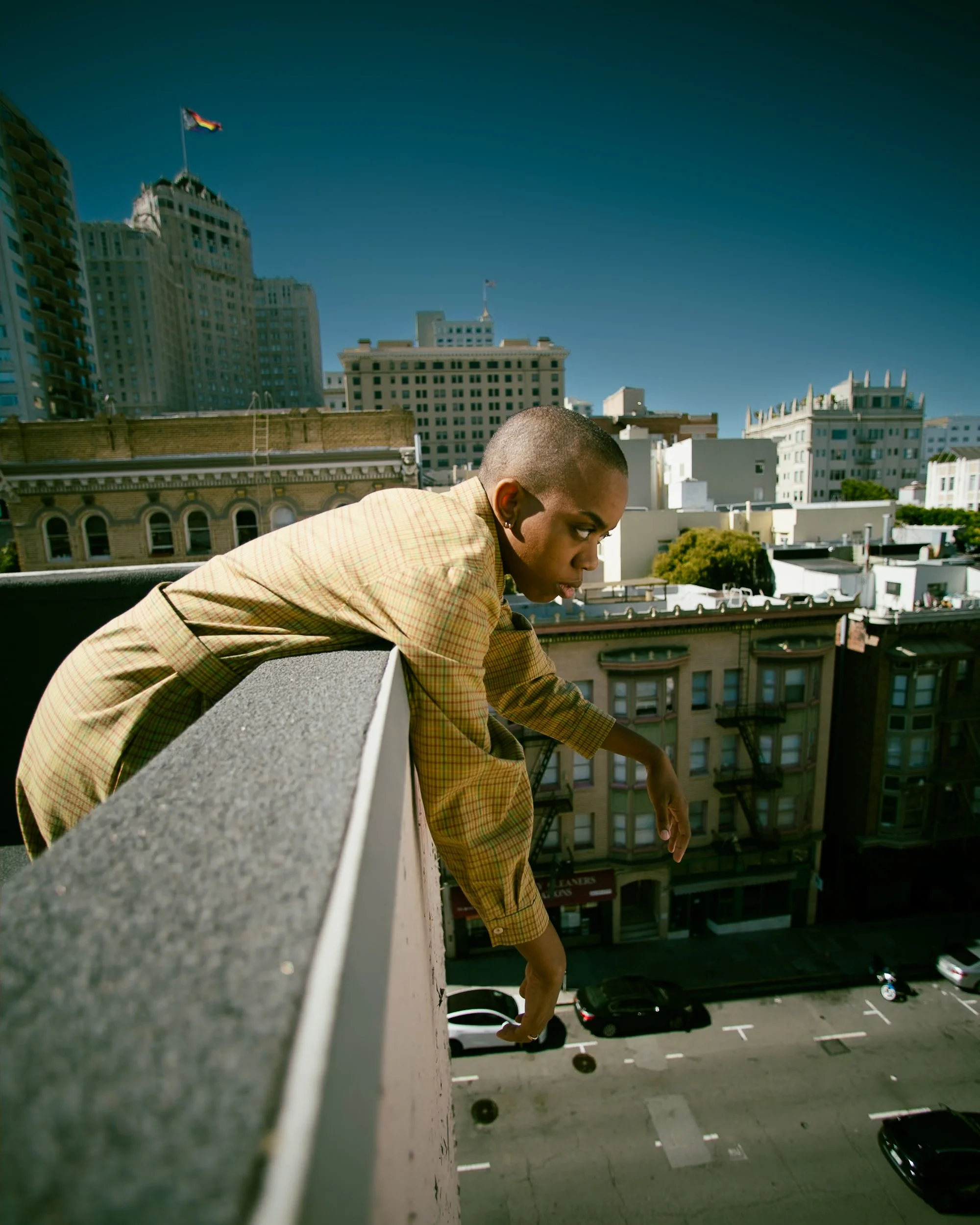 A person leaning over the edge of a rooftop in an urban cityscape with tall buildings, parked cars, and clear blue sky, photographed during a fashion photography session in California for a portrait editorial with fashion photographer..