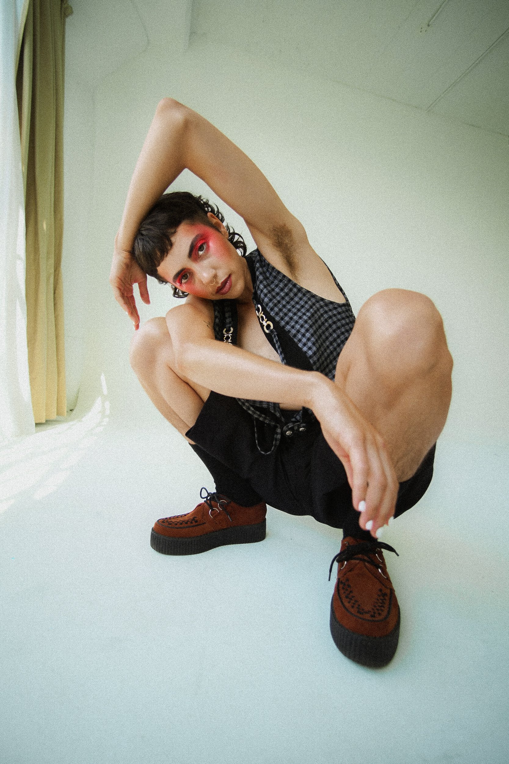 A person wearing a checkered black and blue sleeveless top, black shorts, and brown moccasin-style shoes poses on the floor with one arm over their head and the other hand on their foot, in a room with pale green walls and yellow curtains, photograph