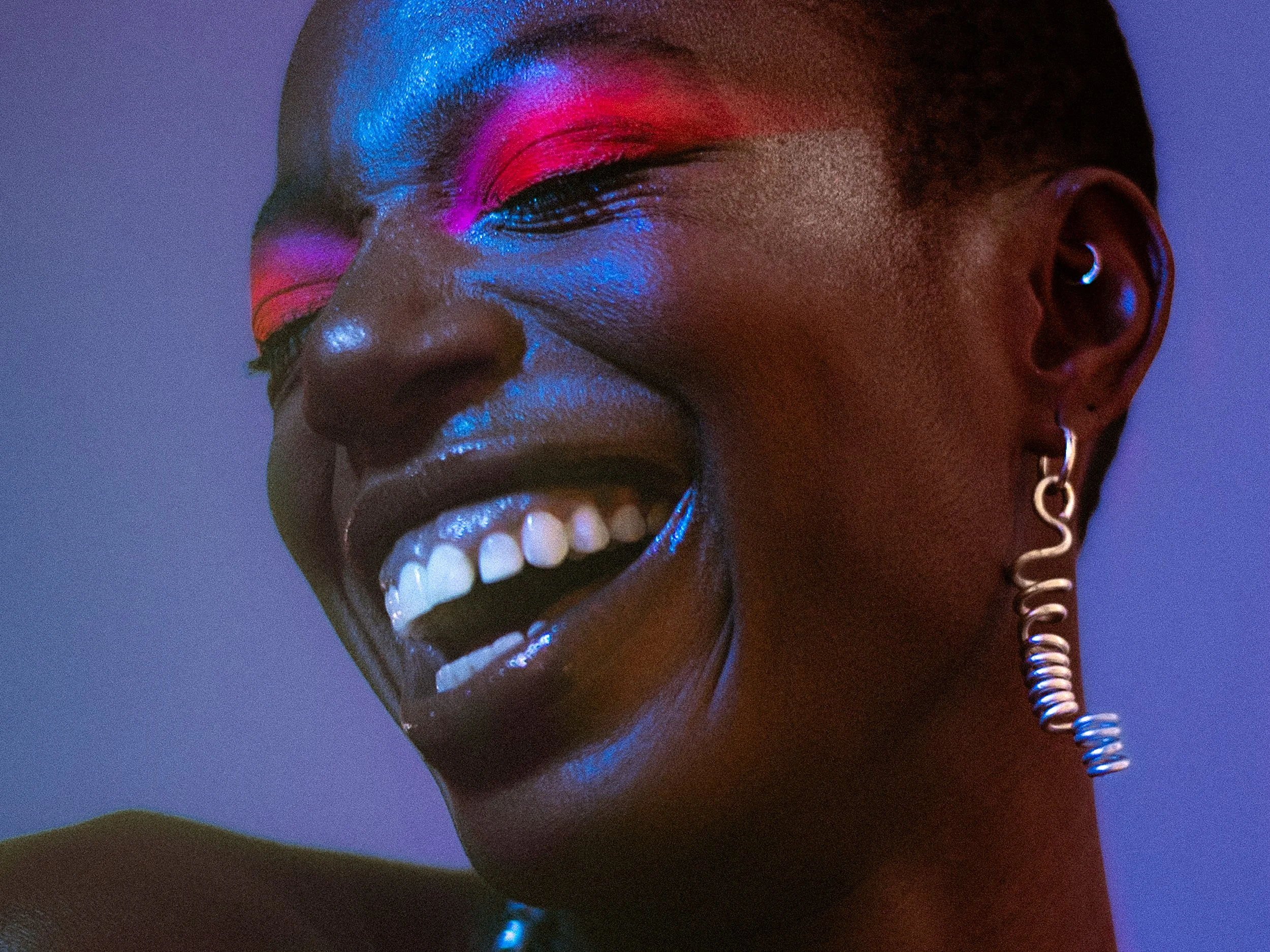 Close-up of a model with dark skin, brightly colored eye shadow, shiny lipstick, and a big smile showing her teeth, wearing unique earrings, with purple and blue lighting,A woman with curly black hair wearing bright makeup, earrings, a rainbow beaded