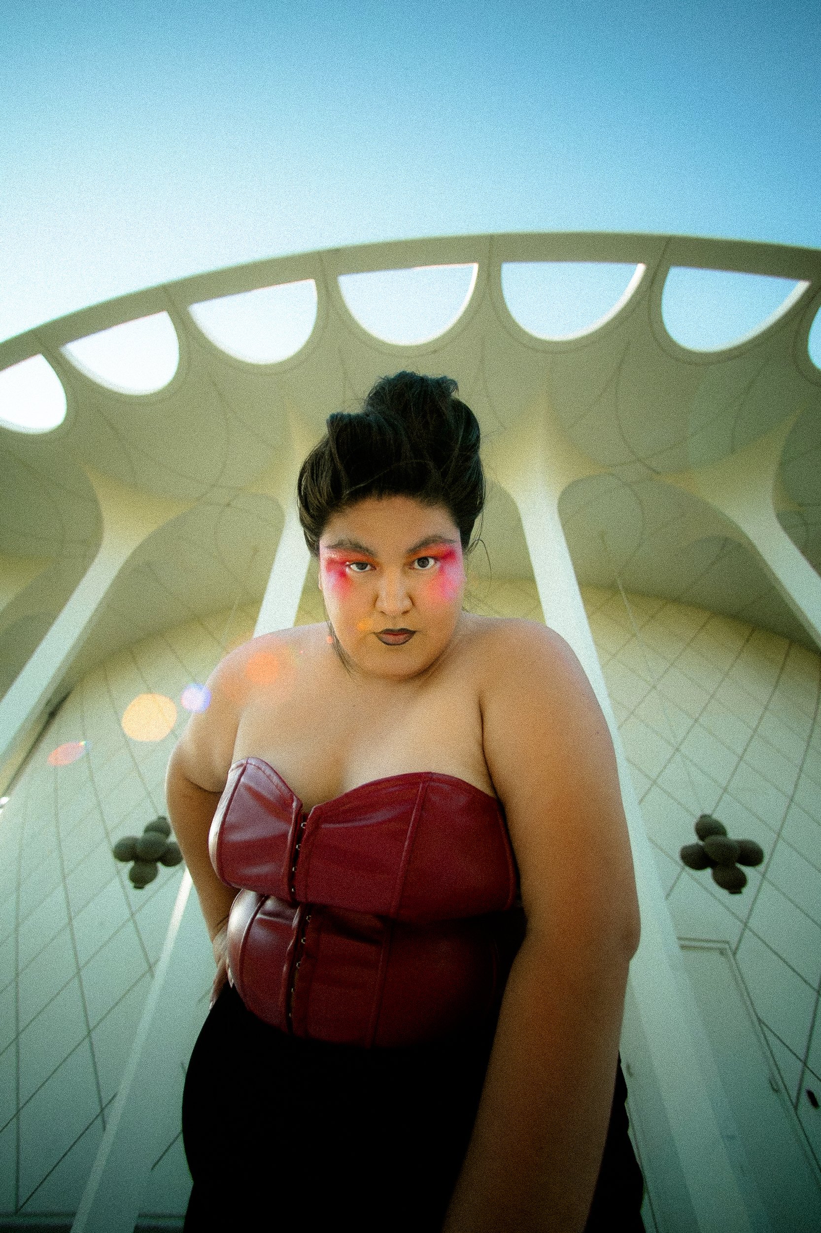 A Latina woman model with bold makeup, dark hair styled up, wearing a strapless red leather top, standing in front of a modern architectural structure with a curved roof and white support beams, taken from a low-angle perspective, photographed during