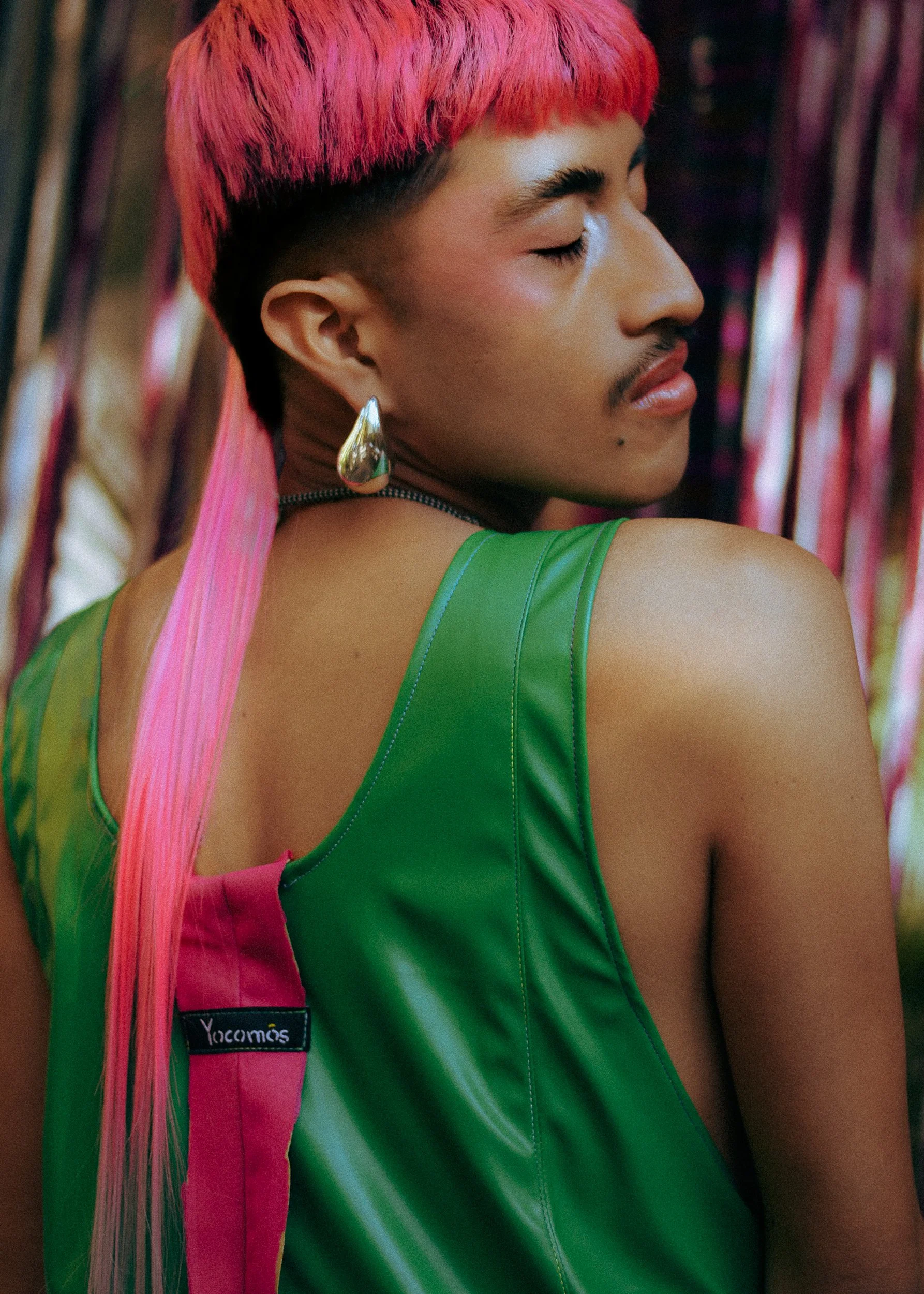 A feminine model with bright pink hair, metallic teardrop earring, wearing a green sleeveless top, with a pink strap bag, and vibrant, multicolored background, photographed during a fashion editorial photography session in Los Angeles with fashion ph