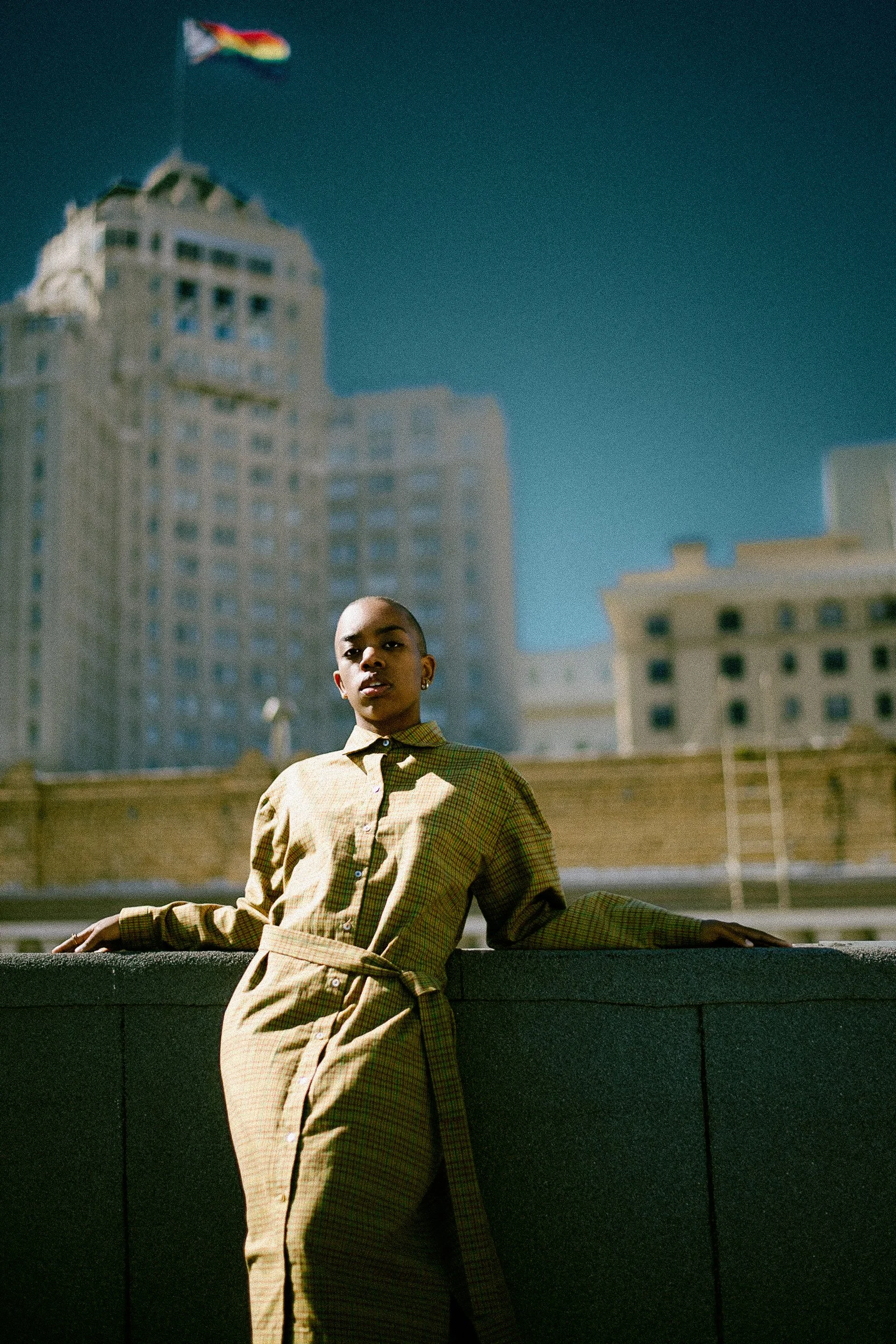 A woman with a shaved head standing outdoors against a cityscape, wearing a yellowish plaid dress with a belt, leaning on a ledge, with tall buildings and a blue sky in the background, photographed during a photography session in California for a por