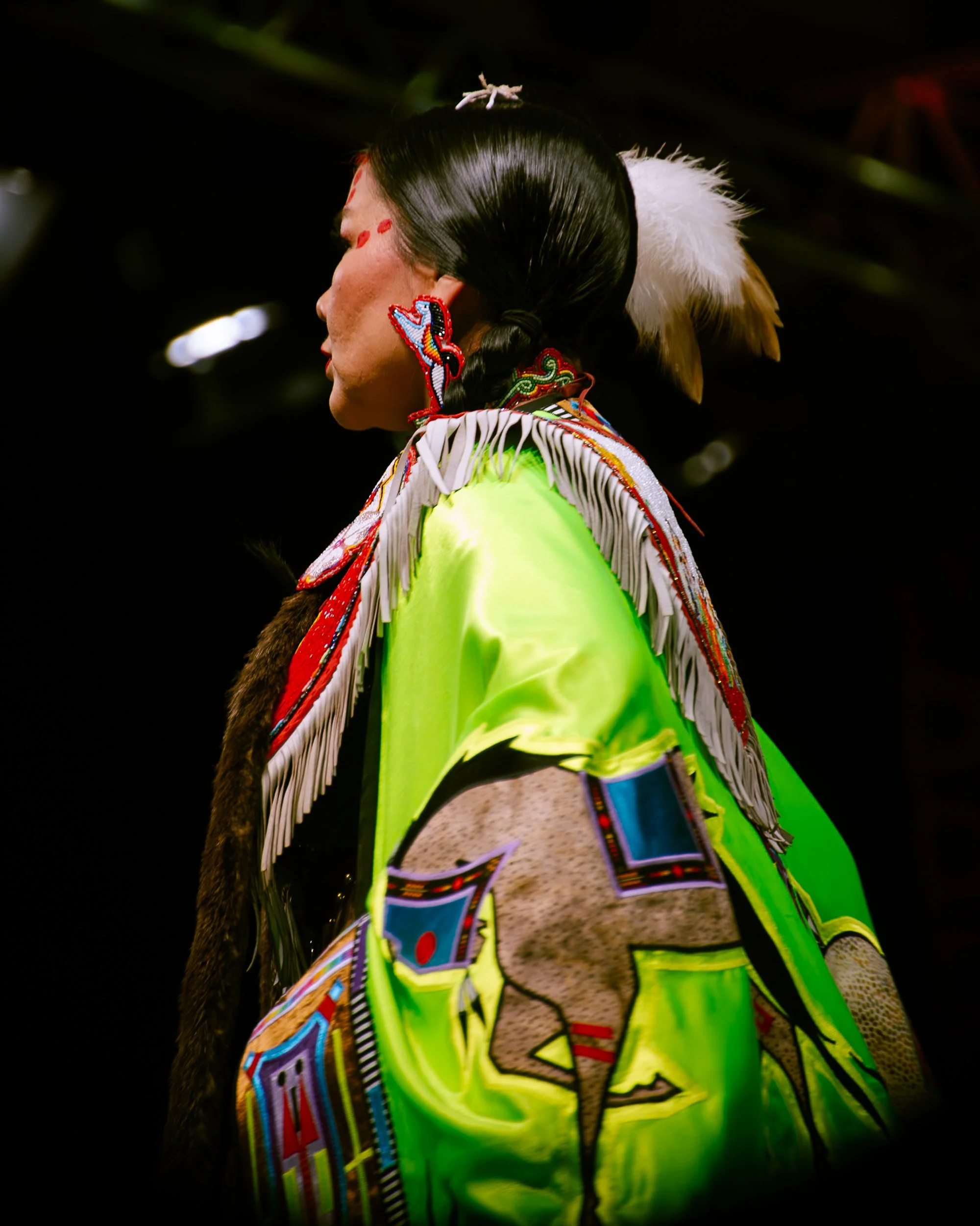 A person in traditional indigenous clothing with colorful beads, feathers, and bright fabric, with a dark background.