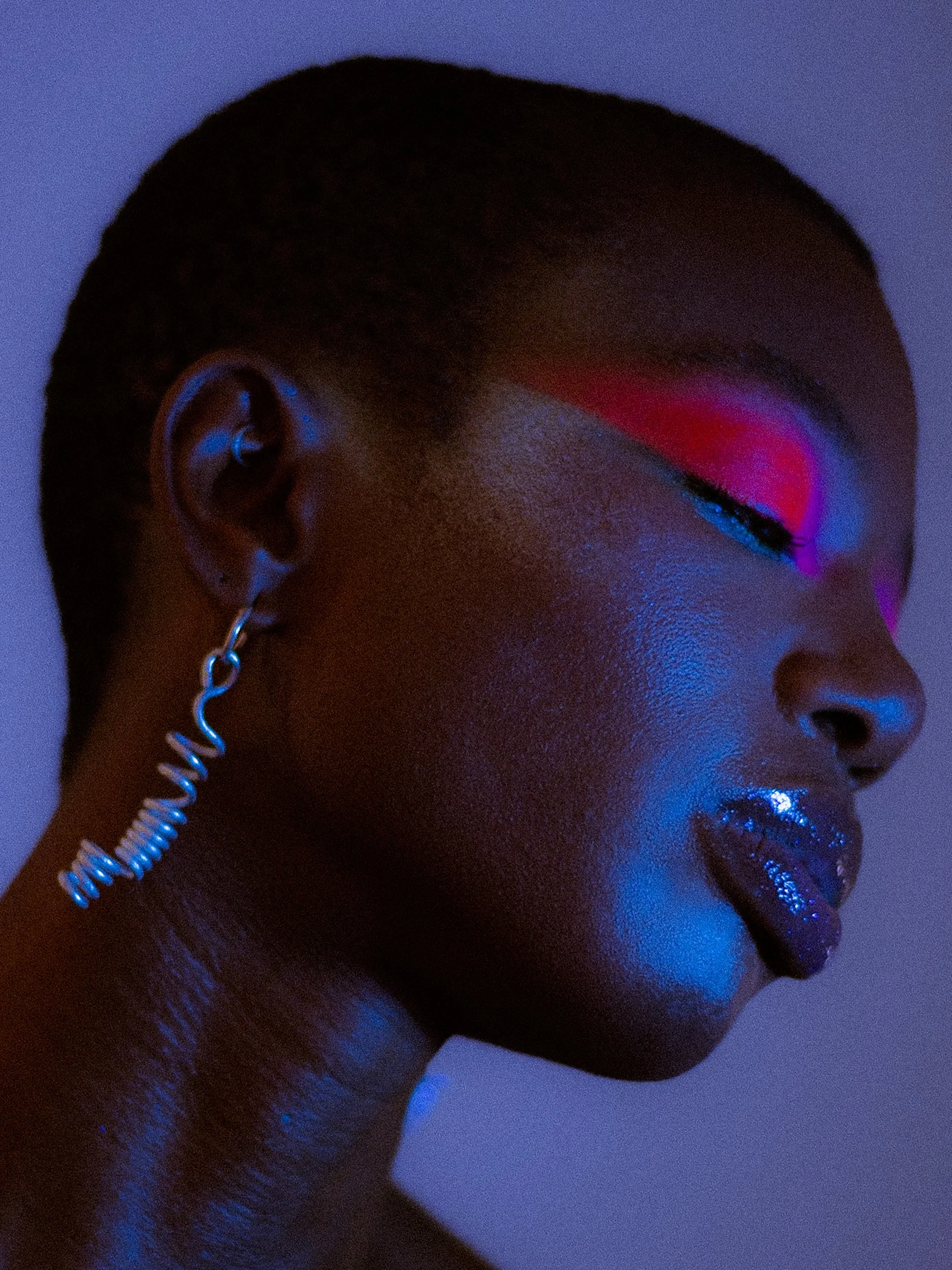 A close-up side profile of a woman model with short hair, closed eyes, and vibrant, colorful makeup illuminated by blue and pink lighting. She is wearing a silver earring, A woman with curly black hair wearing bright makeup, earrings, a rainbow beade
