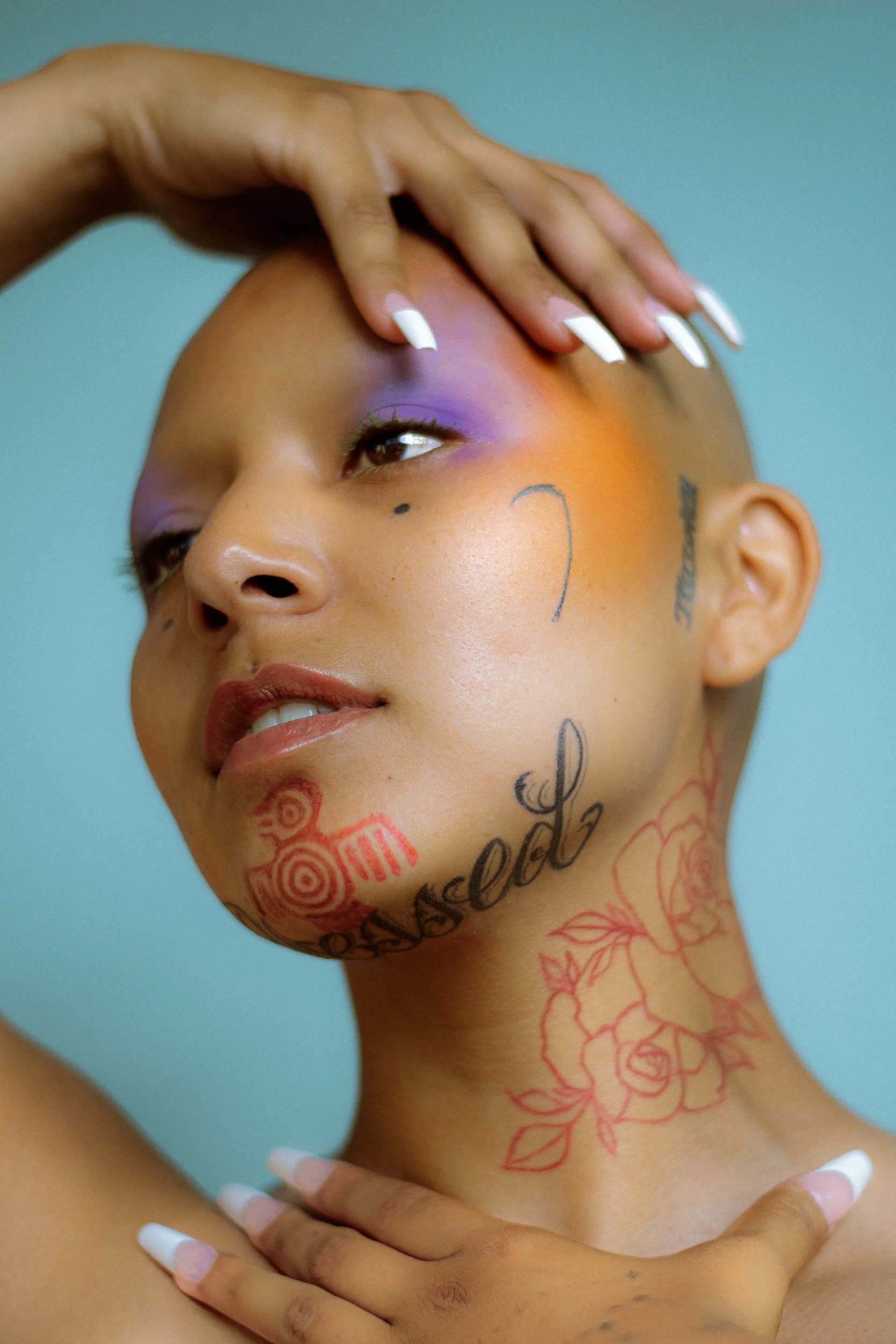 A woman with colorful makeup, tattoos on her face and neck, and white nail polish touches her forehead gently with her hand, photographed during a studio photography session in Los Angeles for a portrait editorial collaboration with fashion photograp
