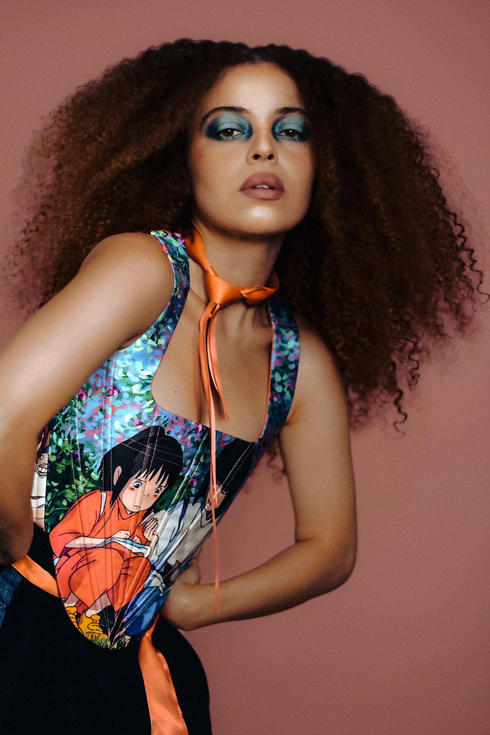 A woman with curly hair and bright blue eyeshadow poses against a pink background, wearing a colorful sleeveless top with a cartoon character design and an orange neck tie, photographed during a fashion editorial photography session in Los Angeles wi