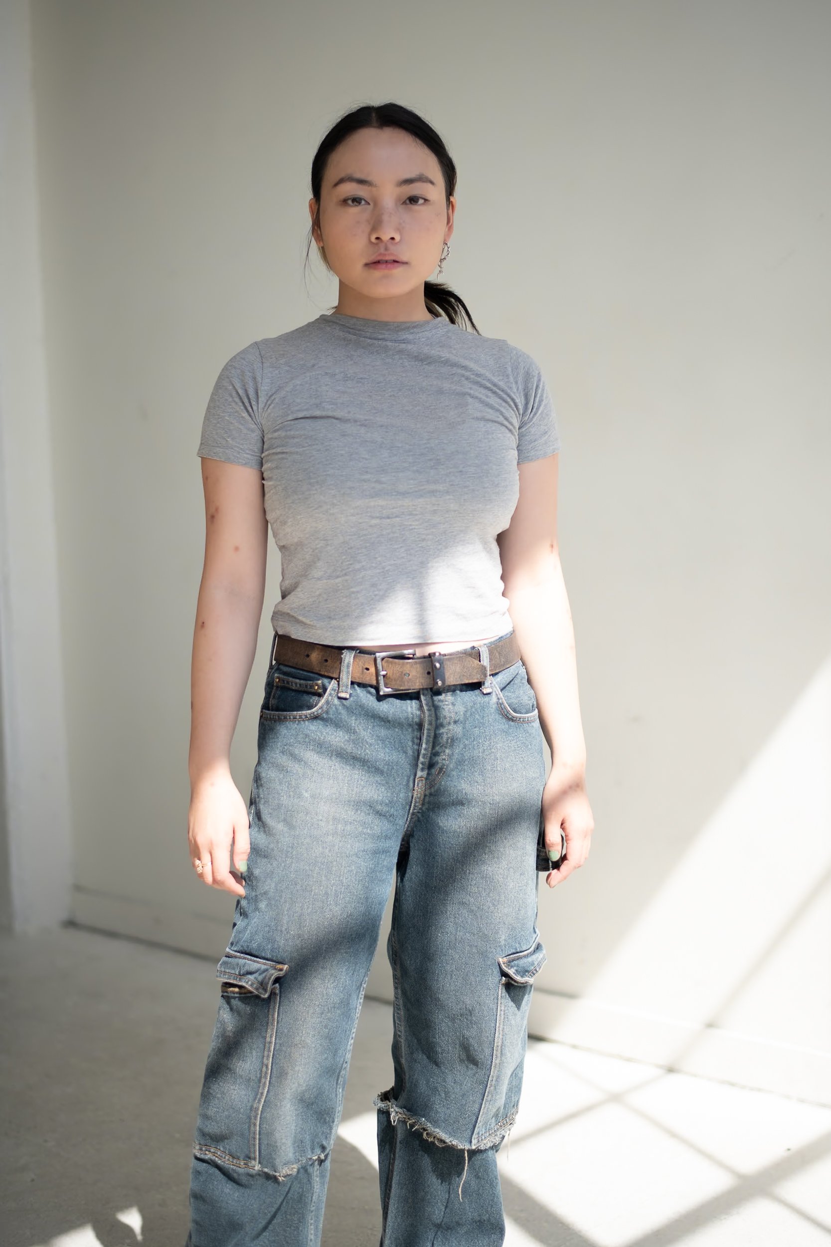 Young woman standing indoors in casual attire, wearing a gray T-shirt and ripped jeans, with natural light coming from the side, photographed during a portrait photography session in Los Angeles with fashion photographer Gí Tenamá.