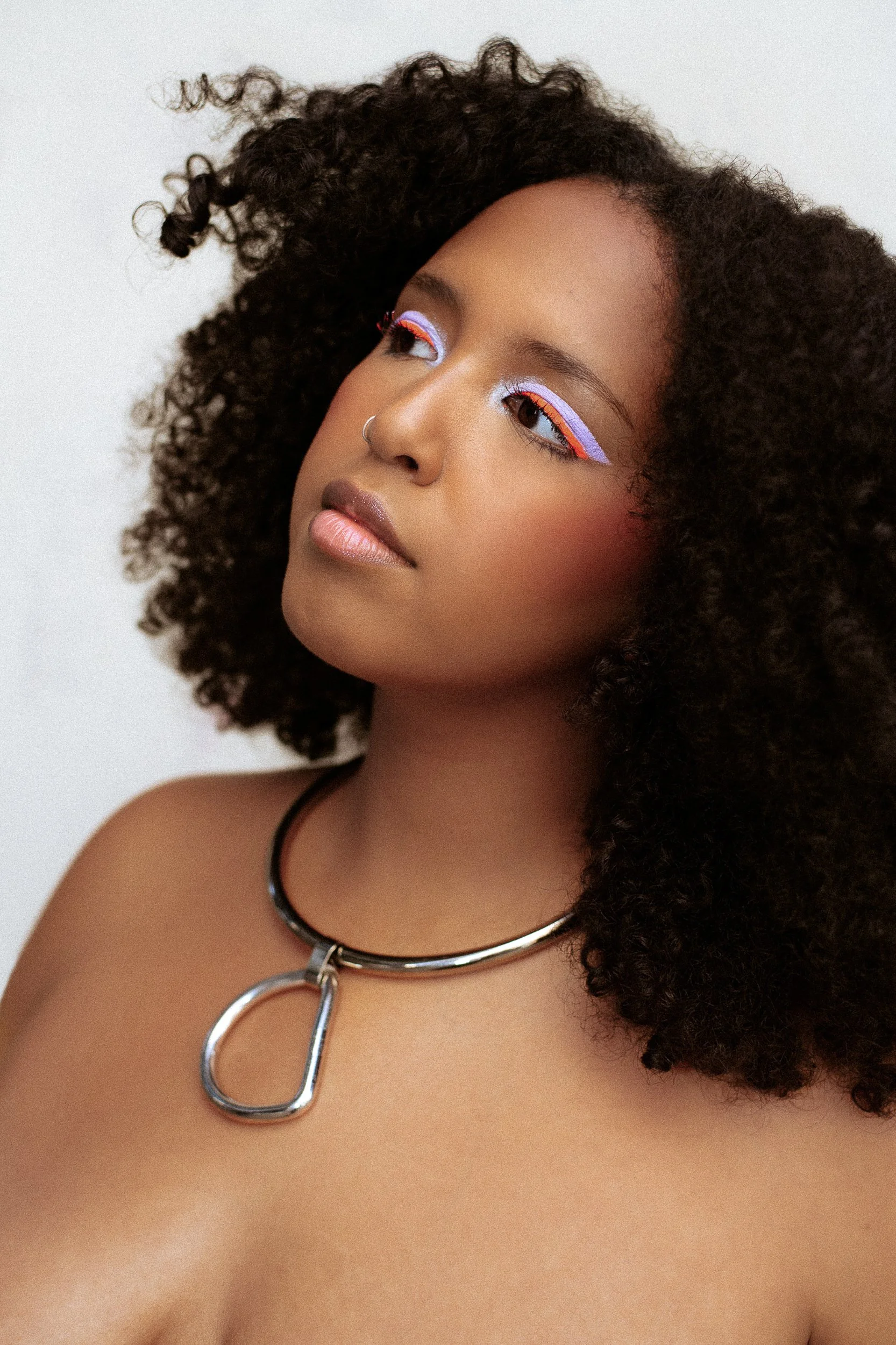 A young woman with curly hair has colorful eye makeup with blue, white, and pink eyeliner, wearing a silver necklace with a large D-shaped pendant, against a plain background, photographed during a studio photography session in Los Angeles for a fash