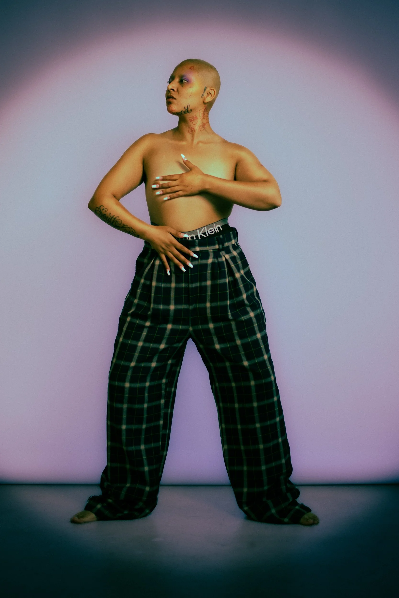 Person with a bald head and tattoos, standing against a plain background, wearing plaid pants with no shirt, with one hand over their chest and the other on their abdomen , photographed during a studio photography session in Los Angeles for a fashion
