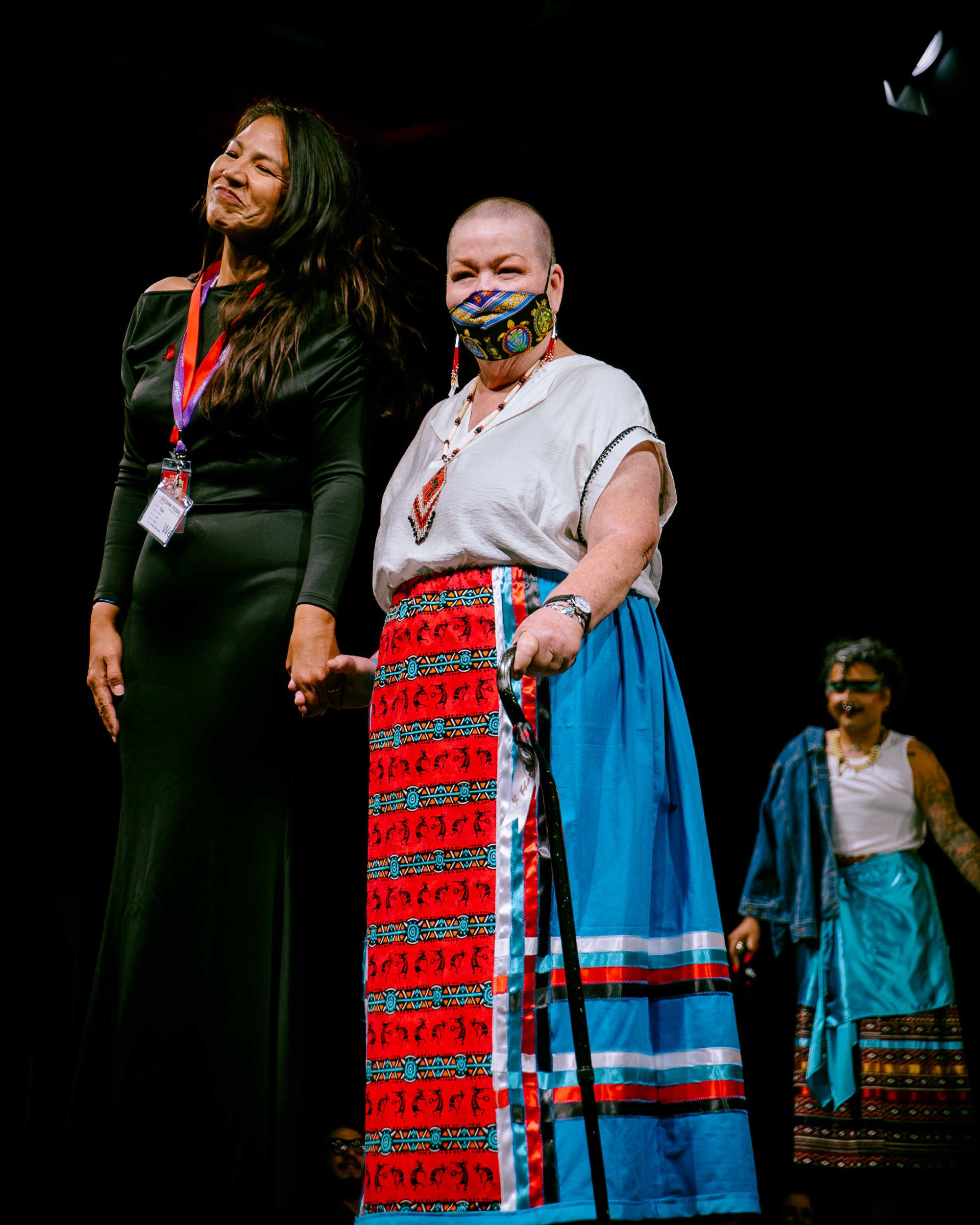 Two women holding hands on stage, one in formal attire and the other in traditional Indigenous clothing, with a woman in the background.