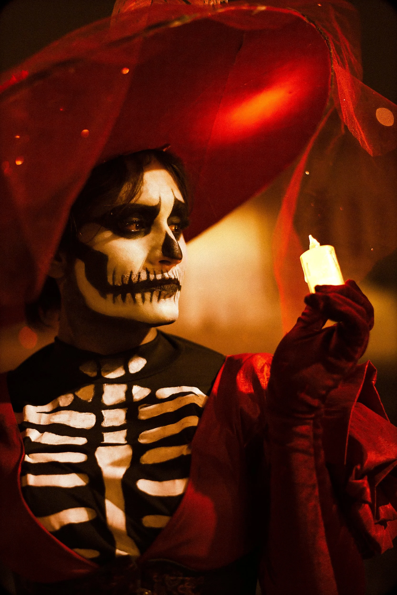 Person dressed in skeleton costume with face painted as skull and black eye makeup, holding a lit candle in a red velvet glove, with a red veil or fabric over their head.
