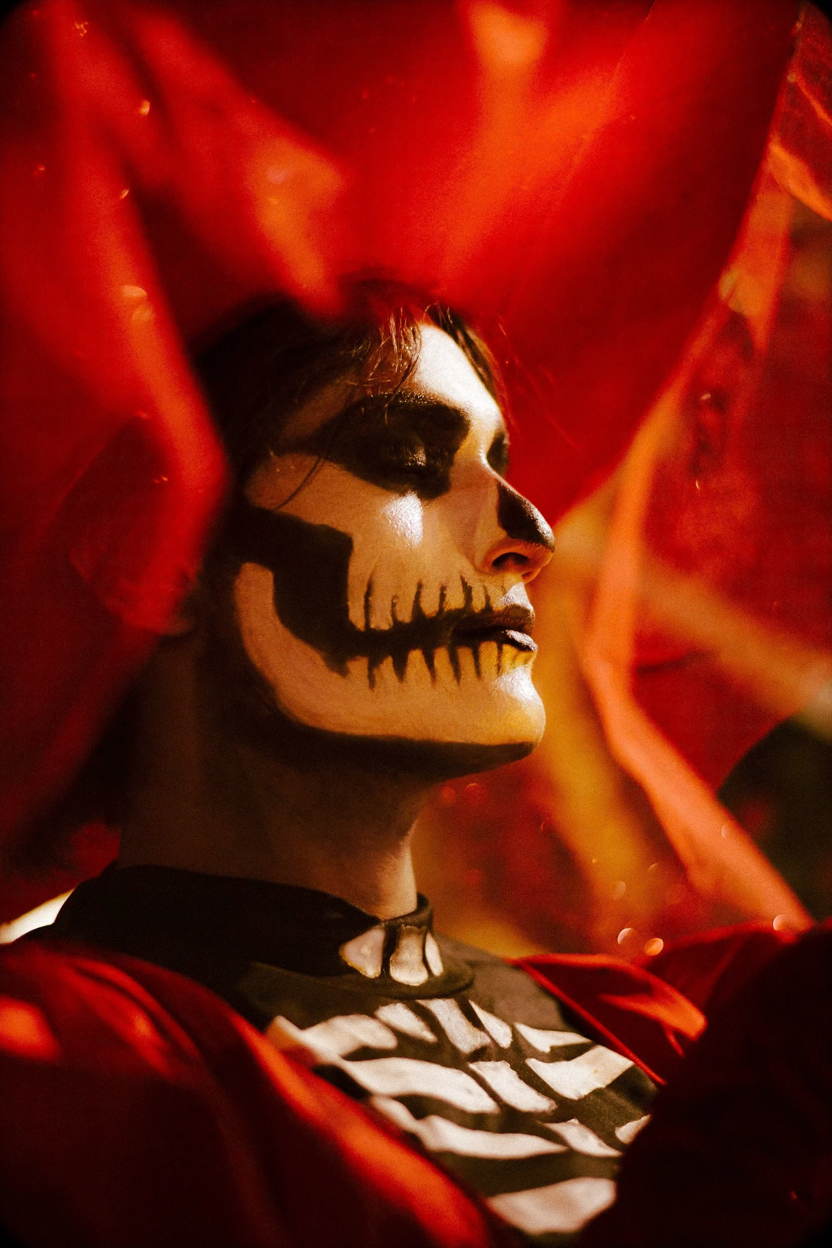 Person with skeletal face paint, wearing a skeleton shirt and red garment, surrounded by orange fabric or decoration.