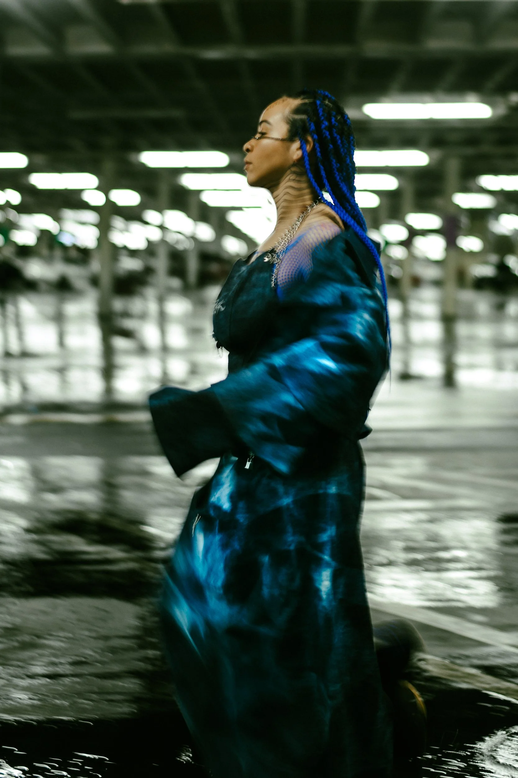 A woman with blue braids, glasses, and layered jewelry walking through an industrial parking garage during the filming of a music video for AnAkA.