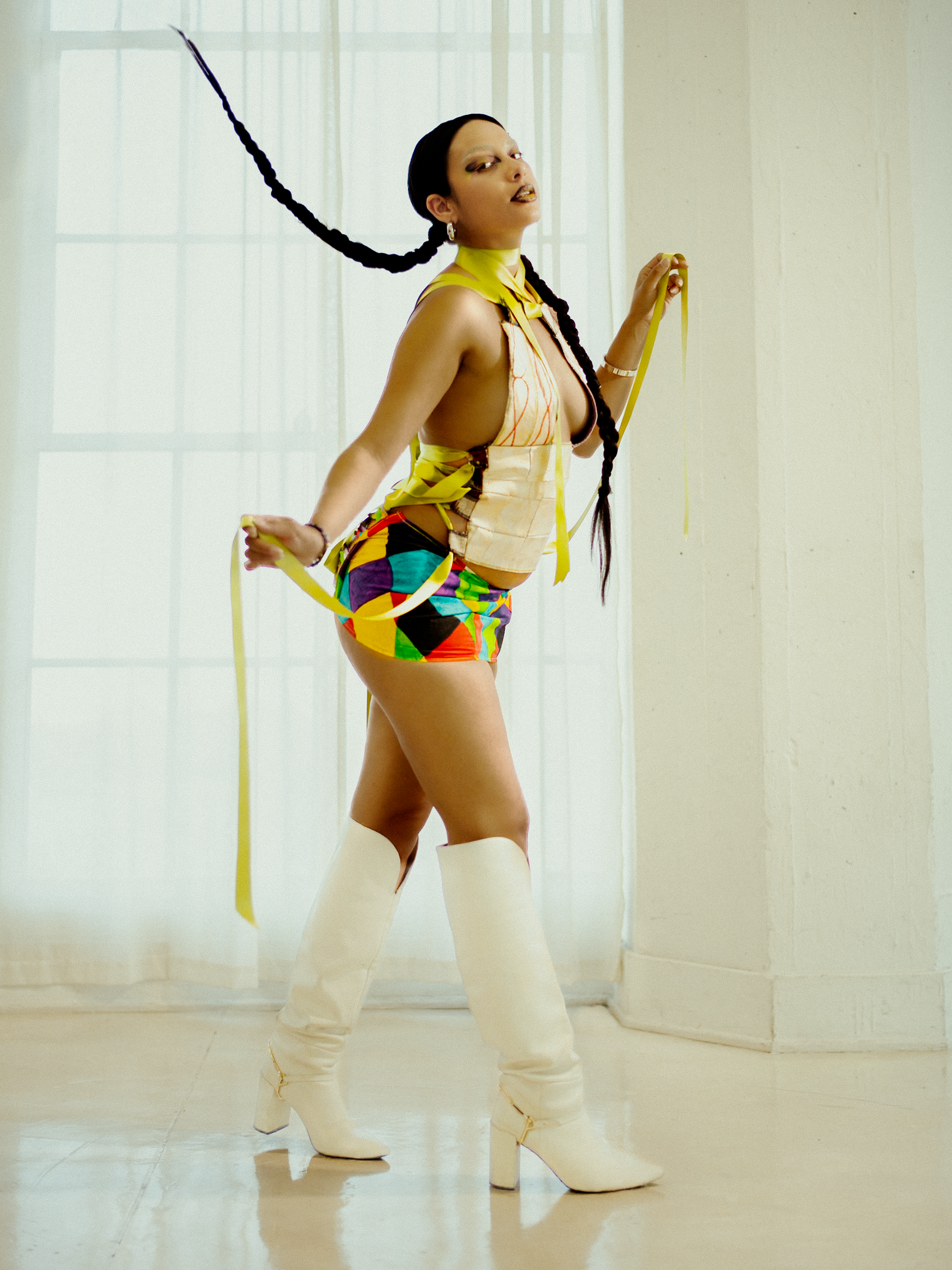 A woman model with long braided hair wearing multicolored outfit, yellow makeup, and white boots, posing indoors against a plain wall. photographed during a studio photography session in Los Angeles for a fashion editorial collaboration with fashion 