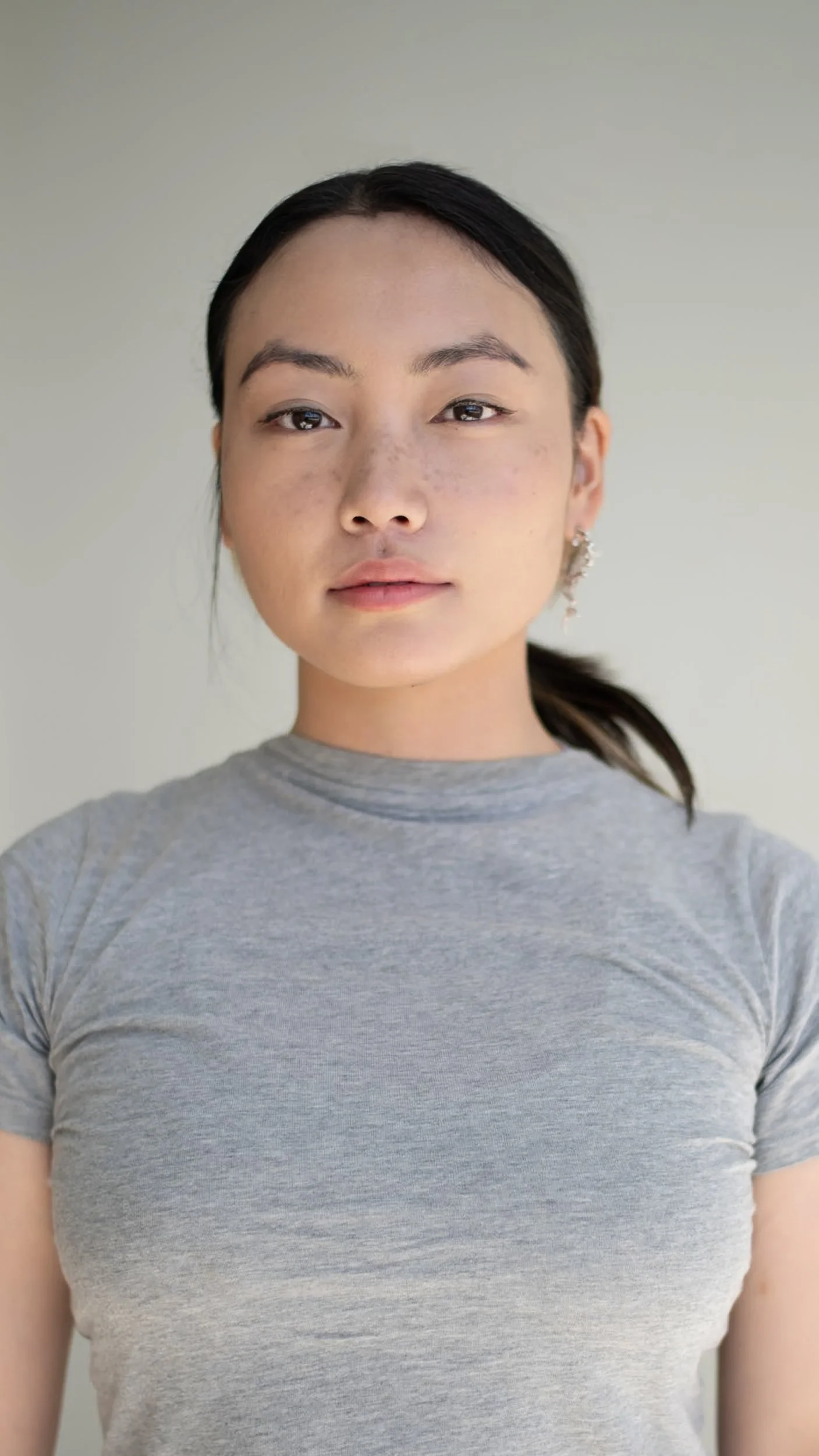 Close-up photo of a young woman with dark hair, wearing a grey t-shirt, standing against a plain background, photographed during a portrait photography session in Los Angeles with fashion photographer Gí Tenamá.