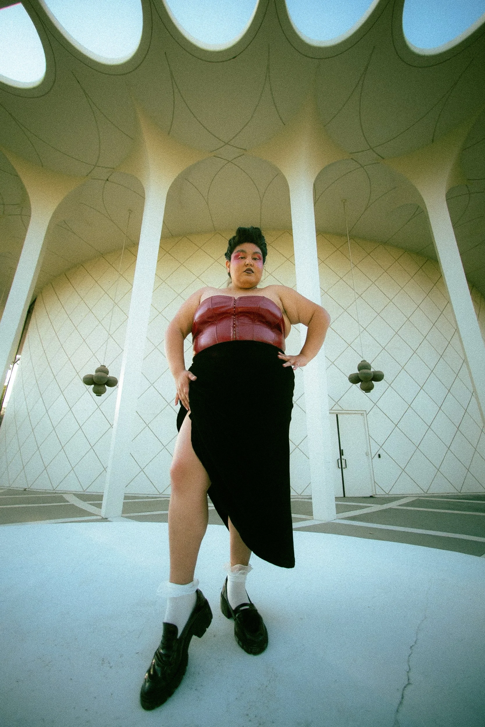 A Latina femme model with short dark hair and colorful makeup wearing a strapless burgundy top, black skirt with a high slit, white socks, and black loafers, standing with hands on hips in front of a modern architectural building with white supports 