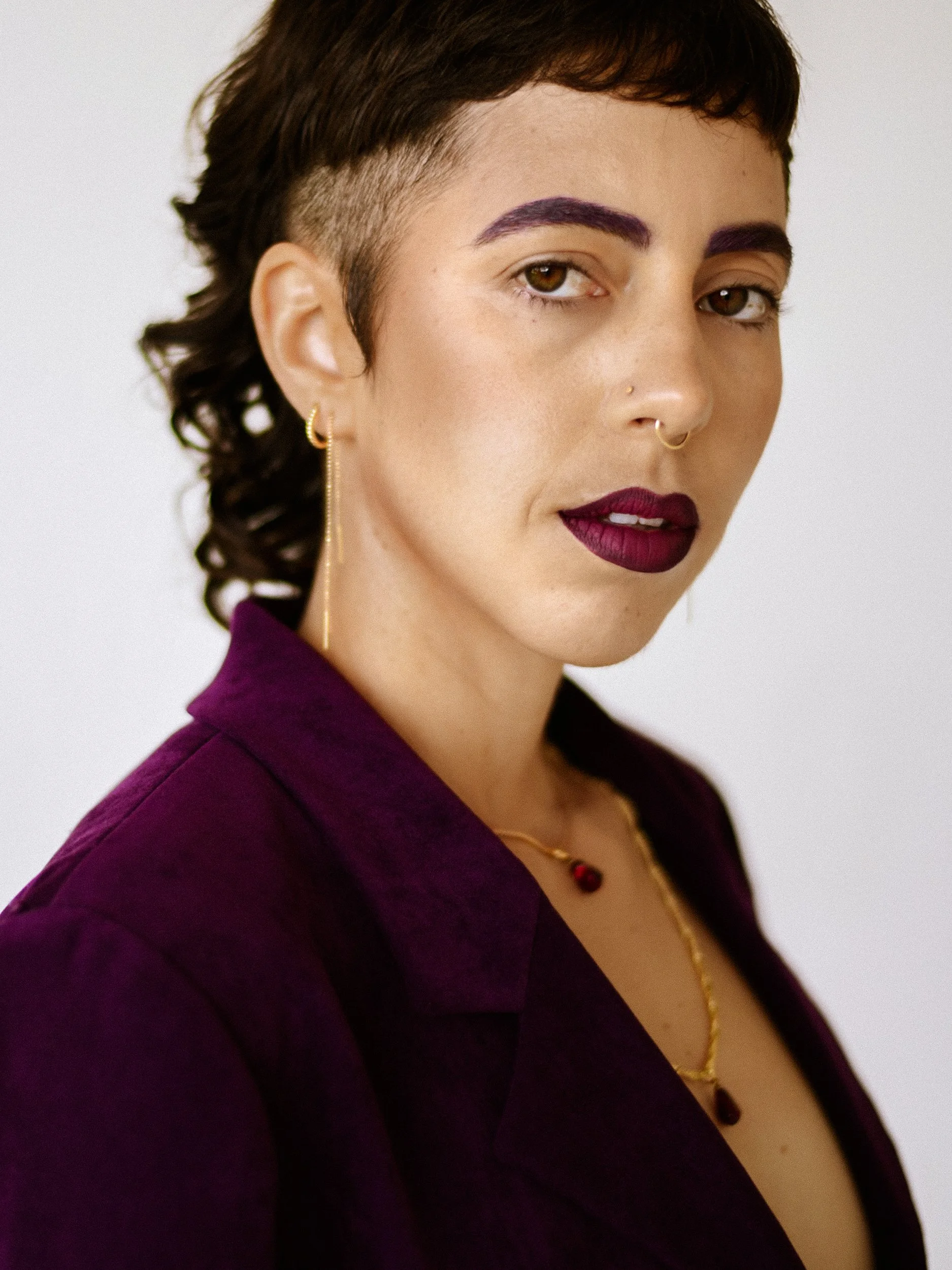 Close-up portrait of a queer nonbinary person with short, dark hair, purple eyebrows, and dark purple lipstick, wearing jewelry including earrings and necklaces, dressed in a dark purple shirt, against a plain white background, photographed during a 