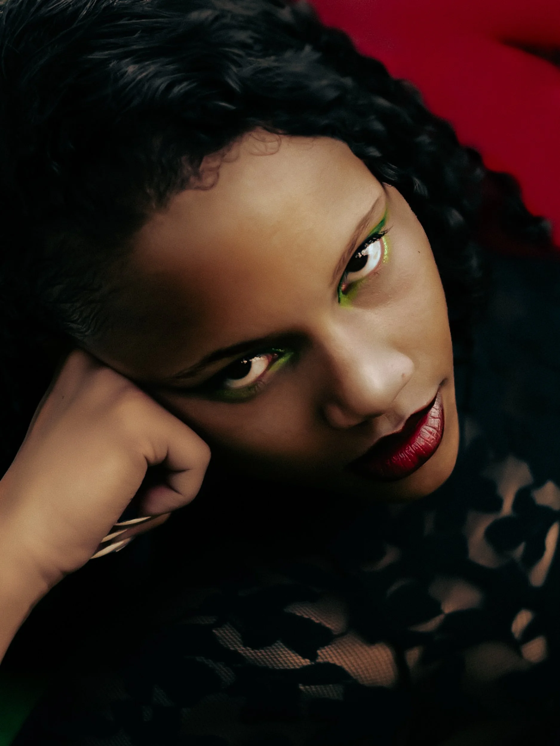 Close-up of a woman model with dark curly hair, green eye makeup, deep red lipstick, and wearing fishnet clothing, resting her head on her hand, photographed during a fashion editorial photography session in Los Angeles with fashion photographer Gí T