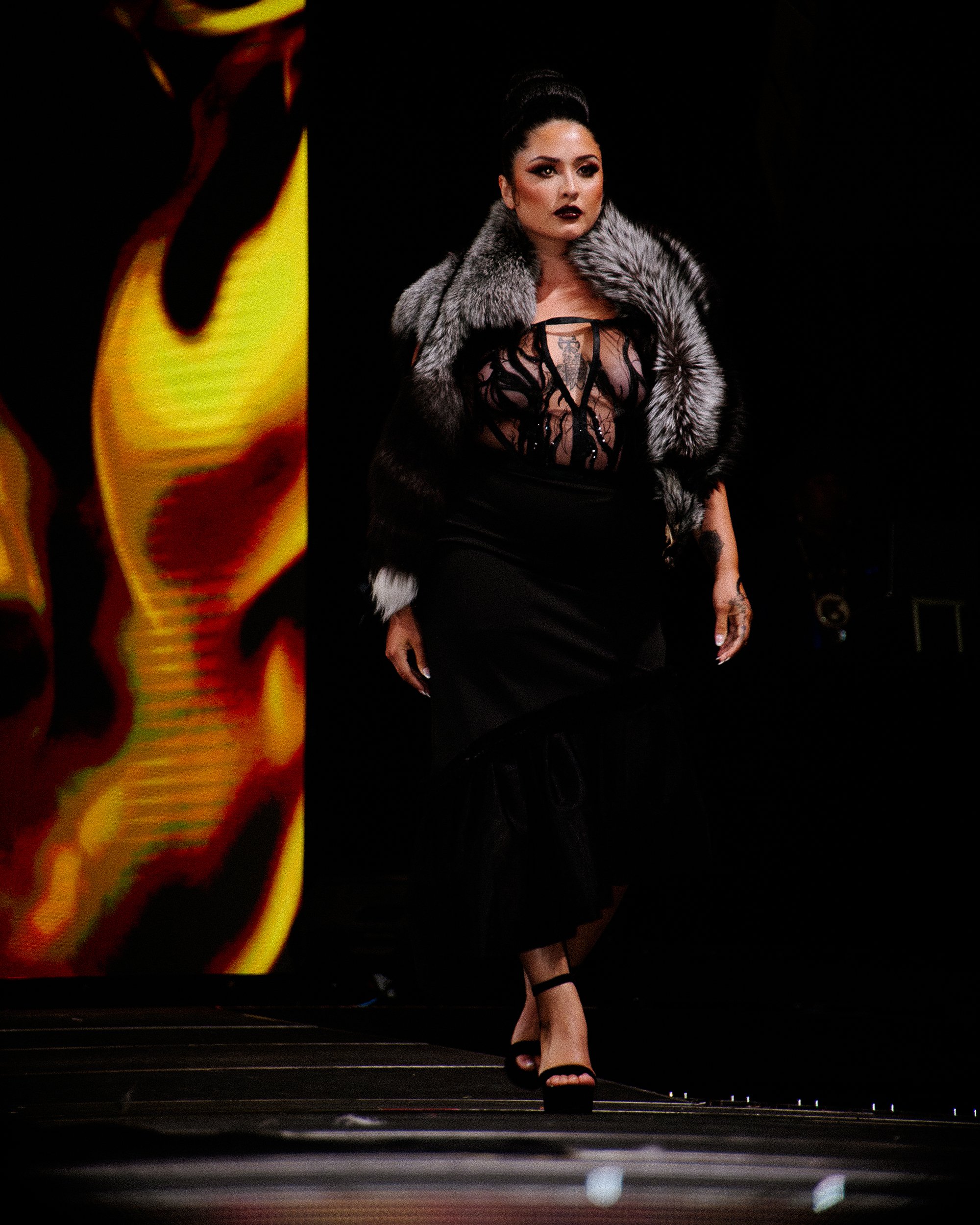 A woman walking on a runway wearing a black dress with sheer embroidery, a fur stole, and high heels.