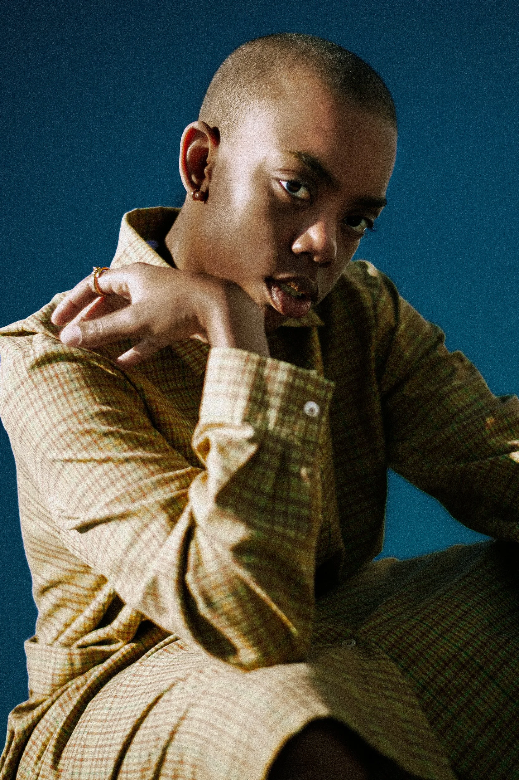 A portrait of a person with a shaved head, wearing a brown plaid shirt, leaning forward with their hand near their face, against a blue background, photographed during a photography session in California for a portrait editorial with fashion photogra