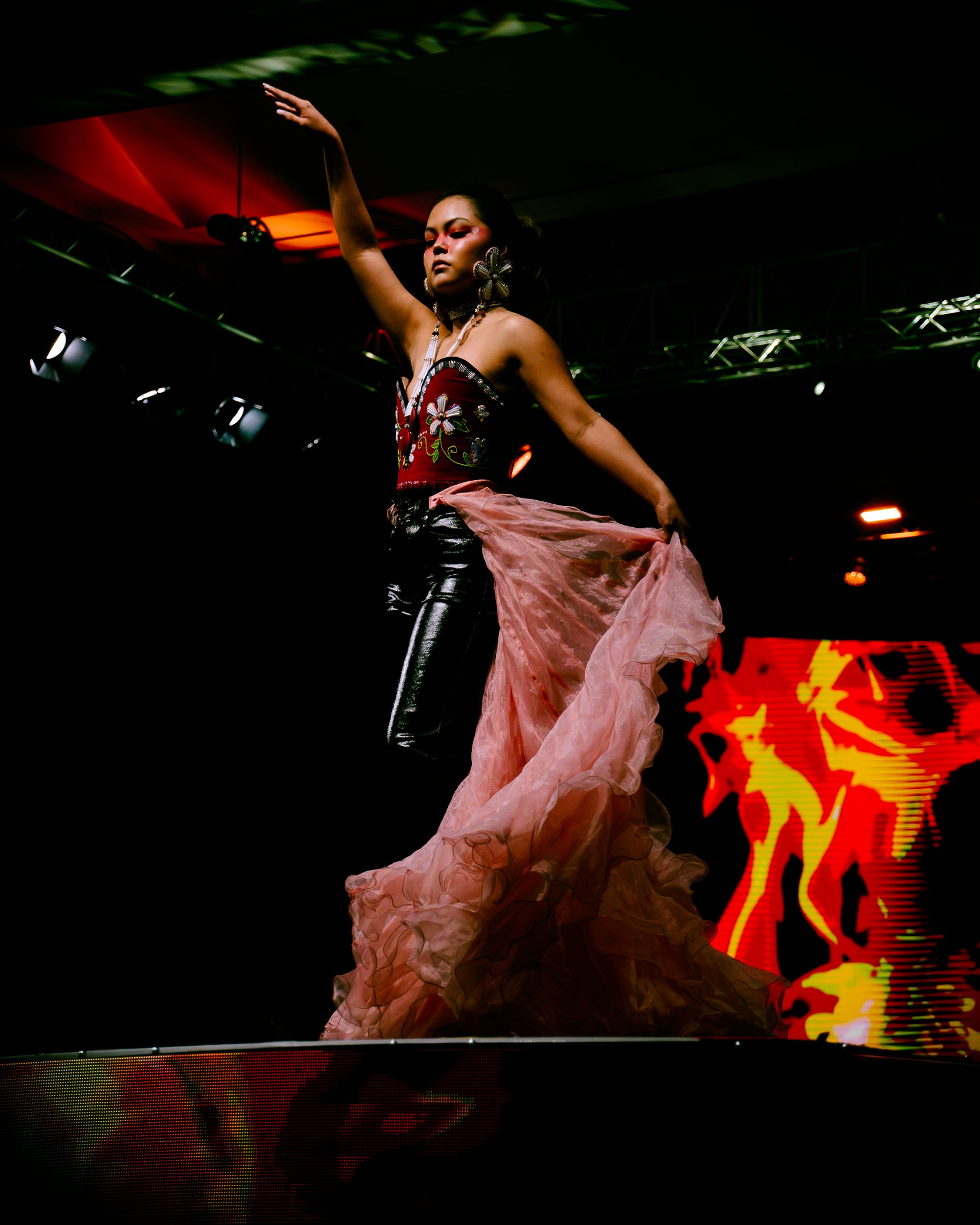 A woman is on stage dancing, wearing a floral embroidered top, shiny black pants, and a flowing pink skirt. She has large earrings and her hair styled back. The stage has colorful lighting and a digital screen with abstract red and yellow visuals in 