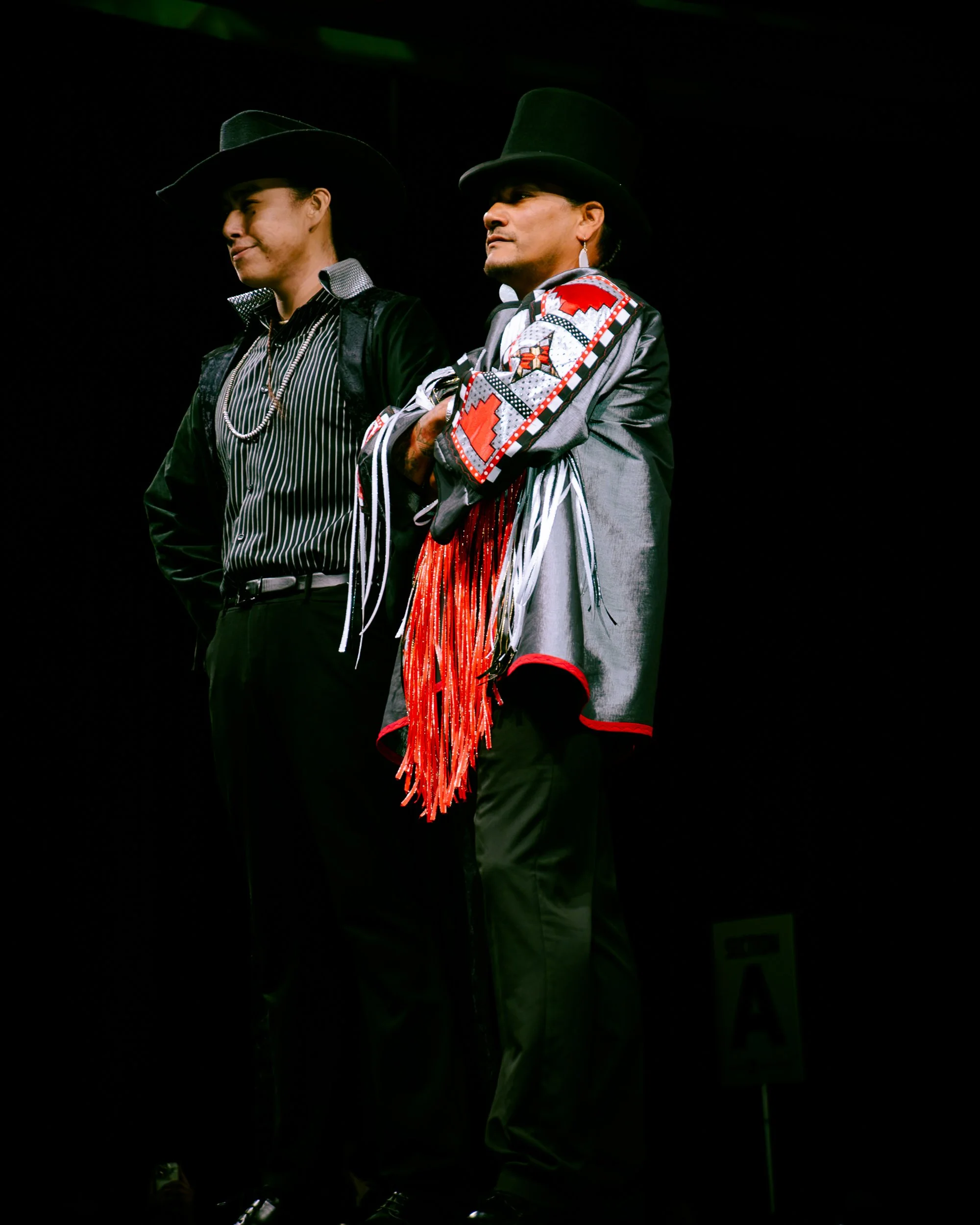 Two individuals on stage wearing traditional Indigenous attire and black hats, standing in a dark setting.