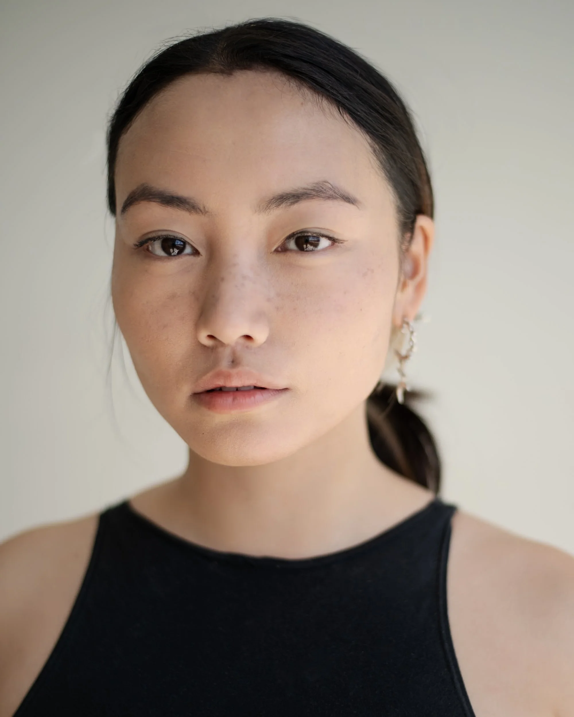 A close-up portrait of a young woman with dark hair, light skin, and freckles, wearing a black sleeveless top and earrings, looking directly at the camera, photographed during a portrait photography session in Los Angeles with fashion photographer Gí