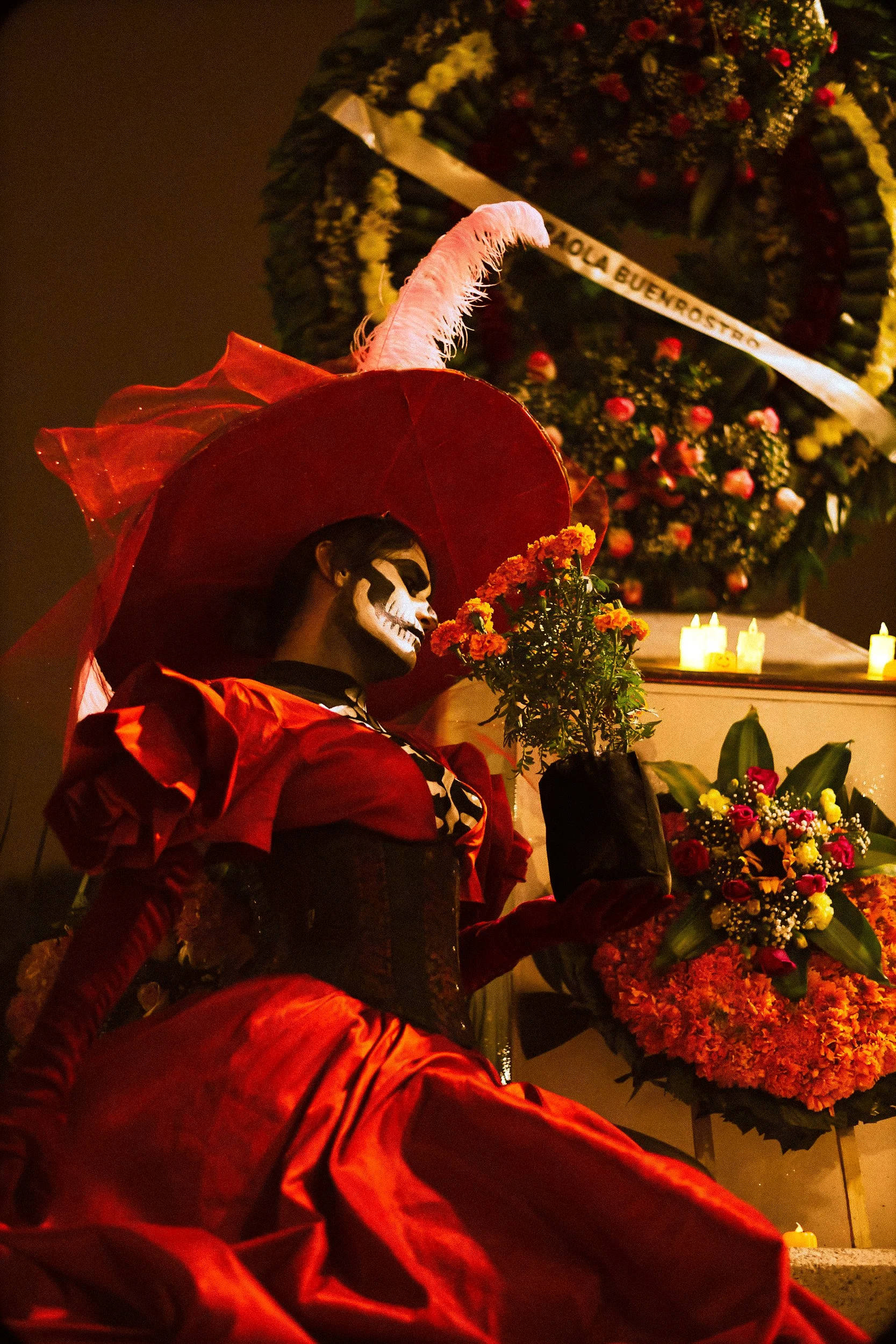 A woman dressed in a red dress with a large red hat with white feather, face painted as a skeleton, holding a potted flower, participating in a Día de los Muertos celebration with candles and flower arrangements in the background.