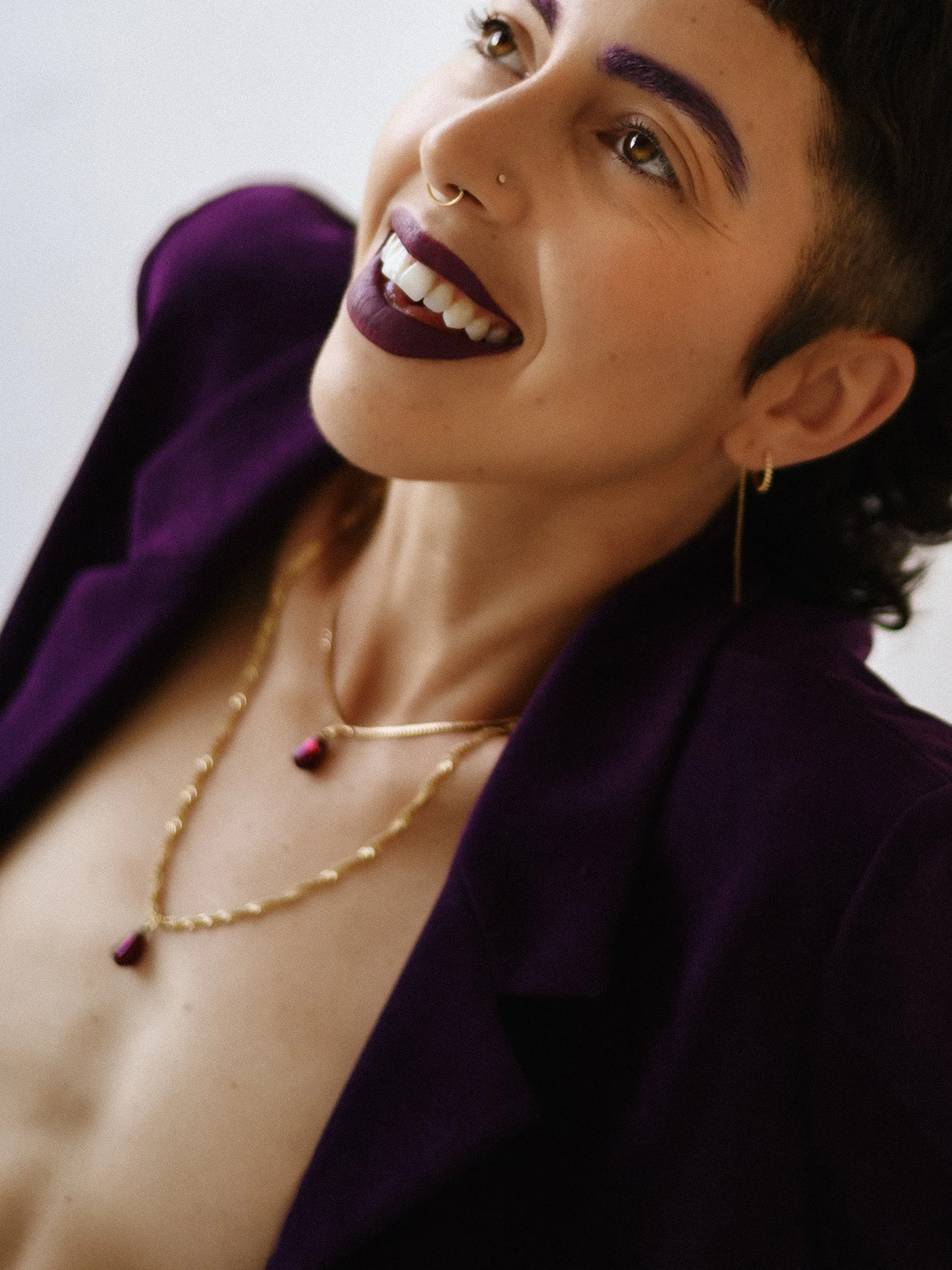Close-up portrait of a queer nonbinary person with short, dark hair, purple eyebrows, and dark purple lipstick, wearing jewelry including earrings and necklaces, dressed in a dark purple shirt, against a plain white background, photographed during a 