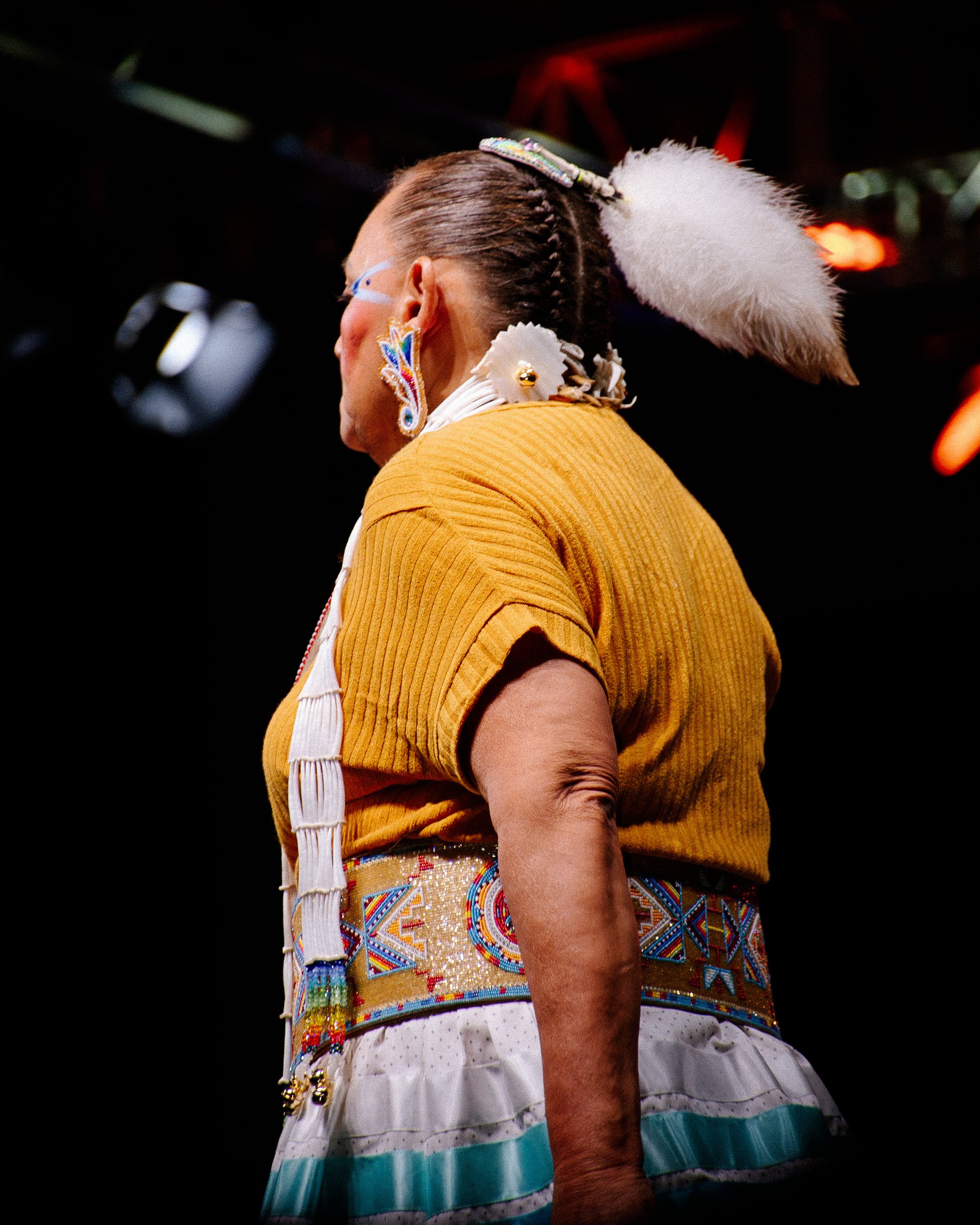 A woman dressed in traditional Native American attire, including a beaded belt, feather headdress, and jewelry, stands in profile against a dark background.