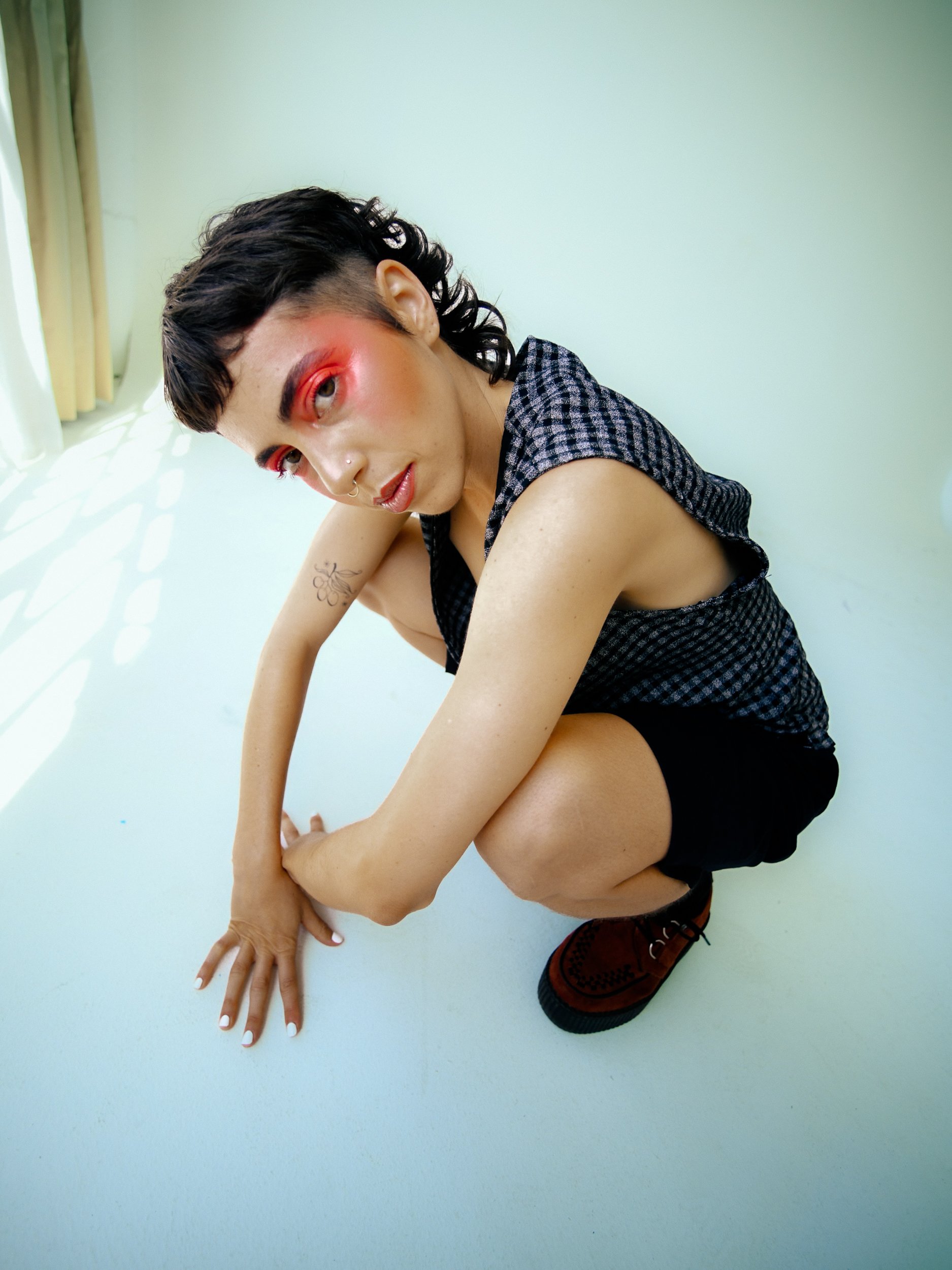 Person crouching on a white floor near a window with light-colored curtains, wearing a checkered sleeveless top and black shorts, with makeup and tattoos., photographed during a studio photography session in Los Angeles for a fashion editorial photos