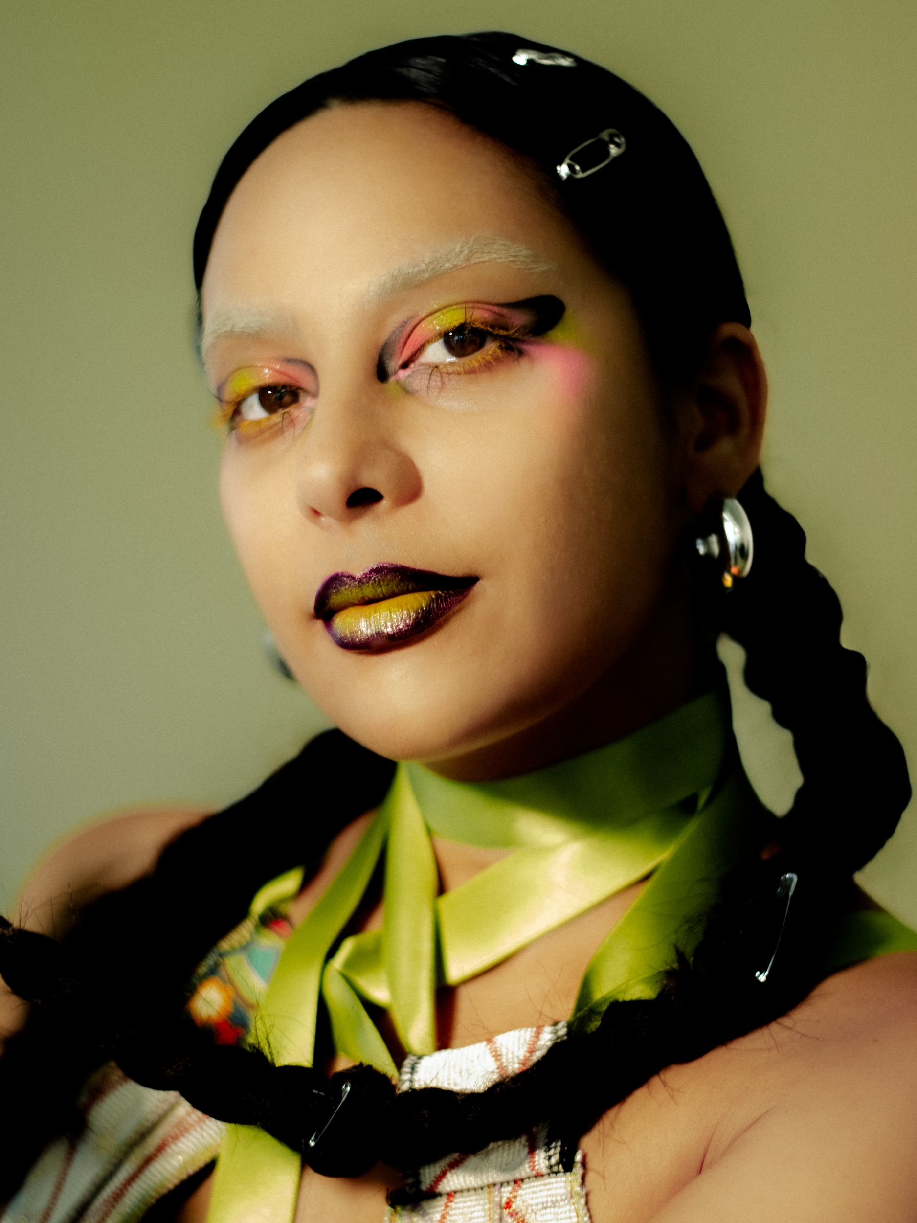 A woman model with long braided hair wearing multicolored outfit, yellow makeup, and white boots, posing indoors against a plain wall. photographed during a studio photography session in Los Angeles for a fashion editorial collaboration with fashion 