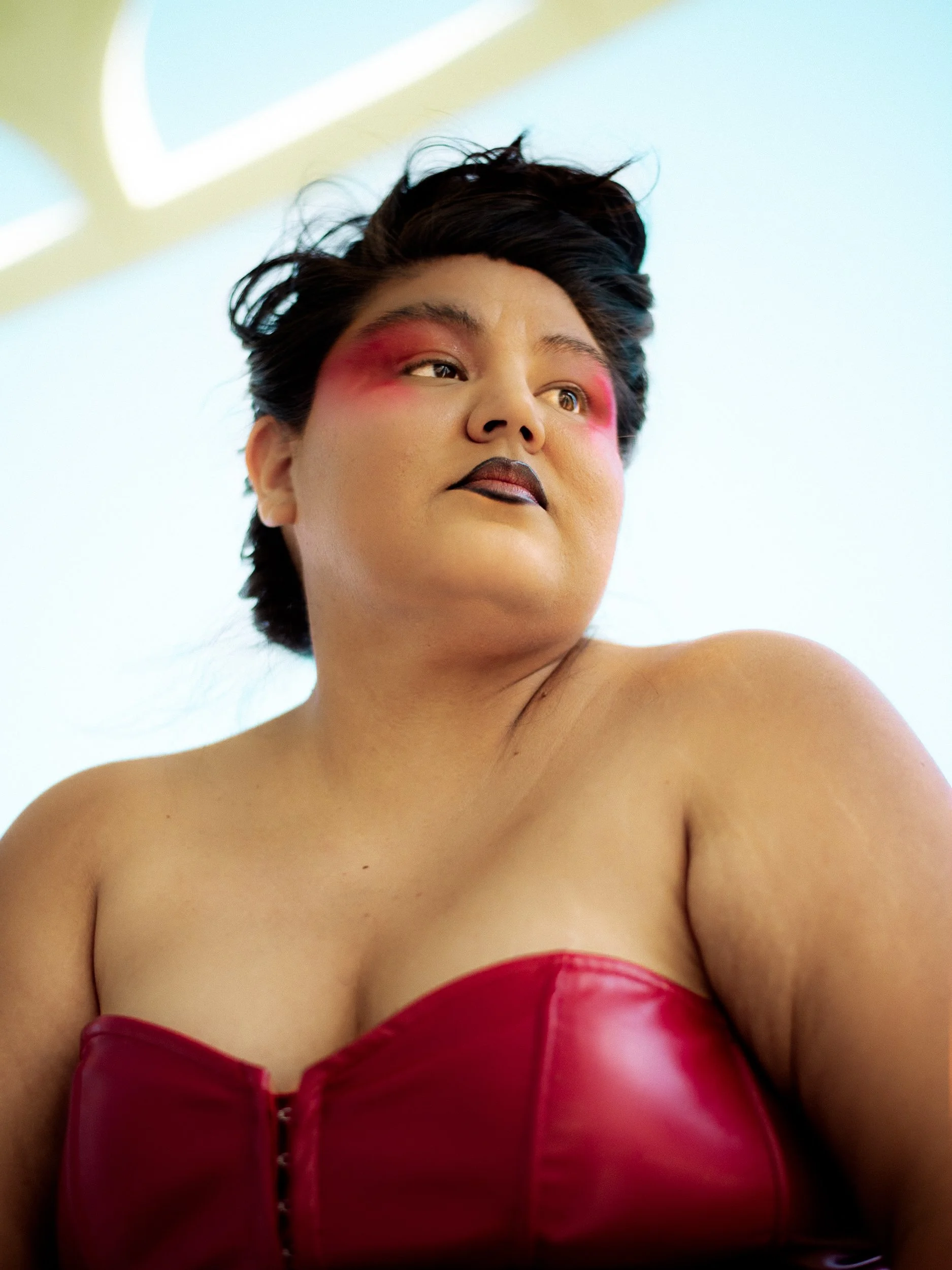 A Latina woman model with short dark hair wearing bold makeup and a red leather strapless top, looking off to the side against a light background, photographed during a fashion editorial photography session in Los Angeles with fashion photographer Gí