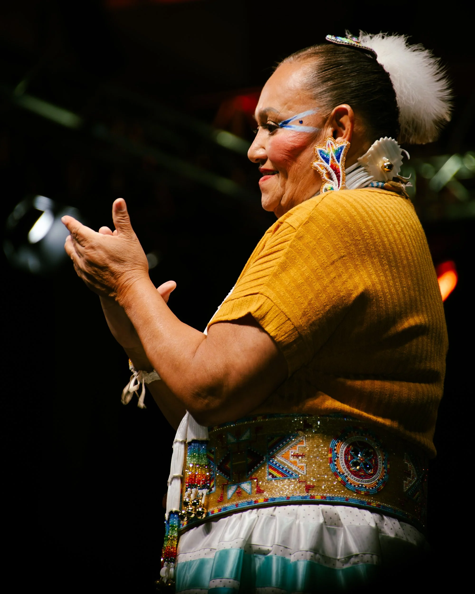 A woman dressed in traditional indigenous attire, clapping and smiling on stage. She wears colorful beaded jewelry, a yellow top, and a beaded belt, with her hair styled back and decorated with feather accents.