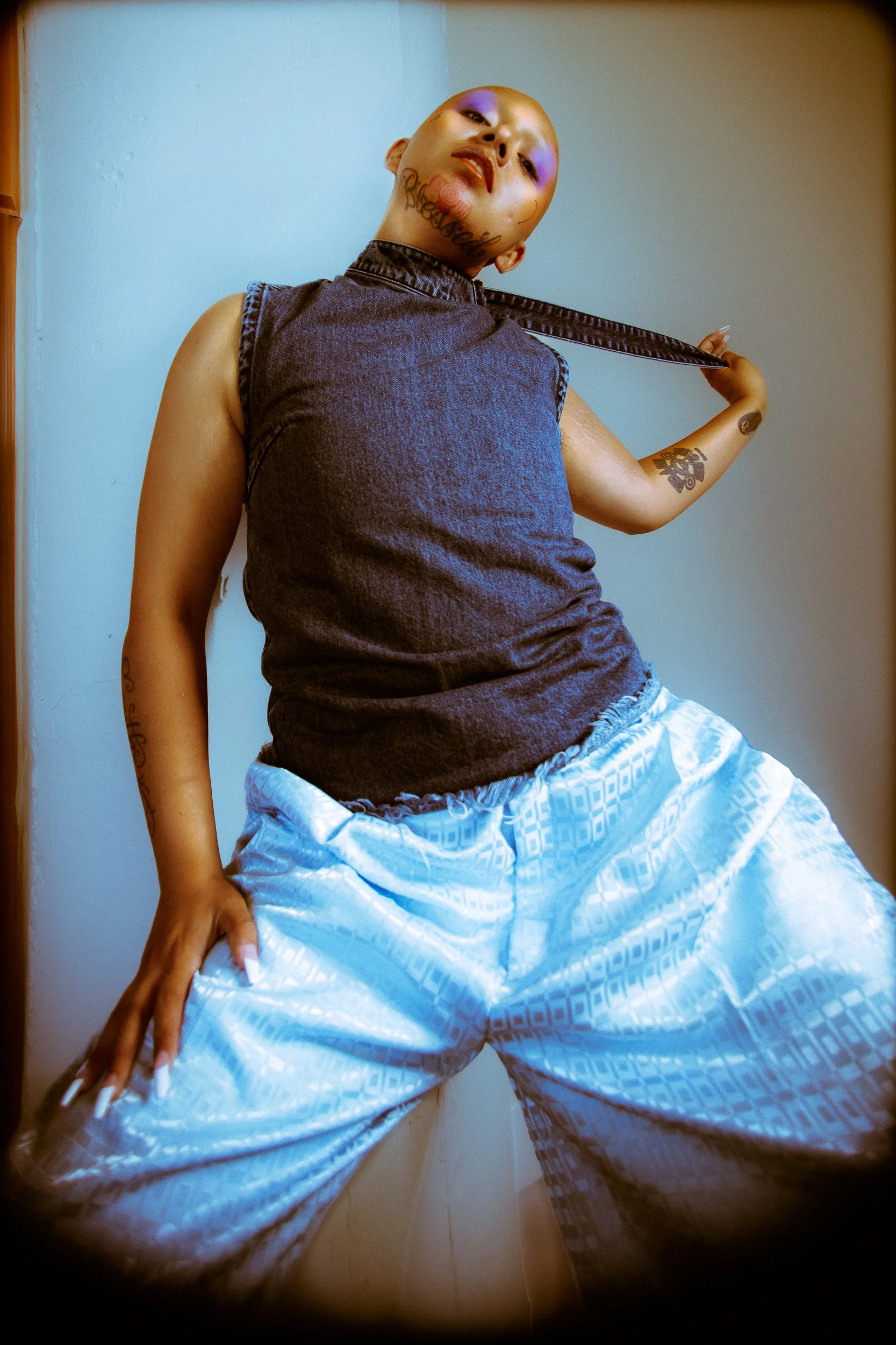 A person with tattoos, wearing a sleeveless denim top and loose patterned light blue pants, poses confidently indoors, holding a strap over their shoulder, photographed during a studio photography session in Los Angeles for a jewelry editorial collab