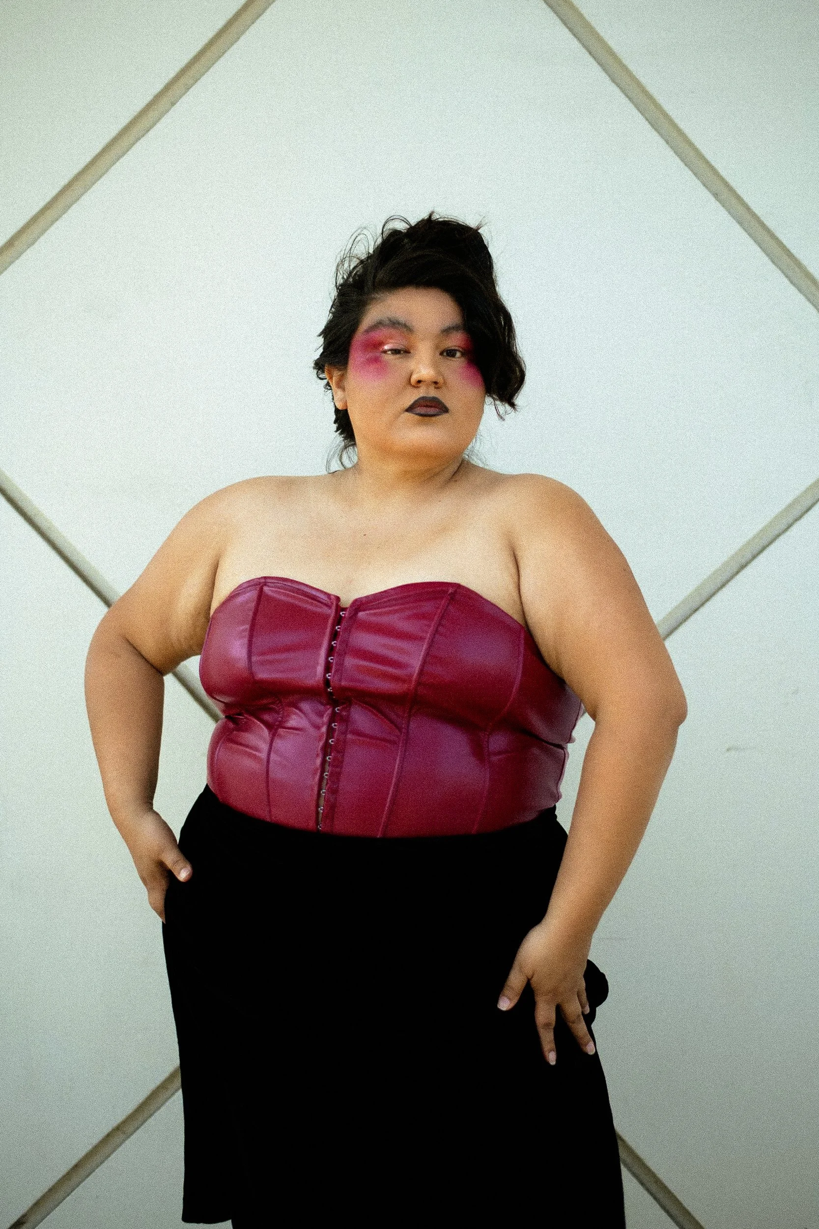 A confident Latina femme model with short dark hair and bold makeup, wearing a strapless burgundy leather corset top and a long black skirt, standing in front of a plain wall with a geometric design background, photographed during a fashion editorial