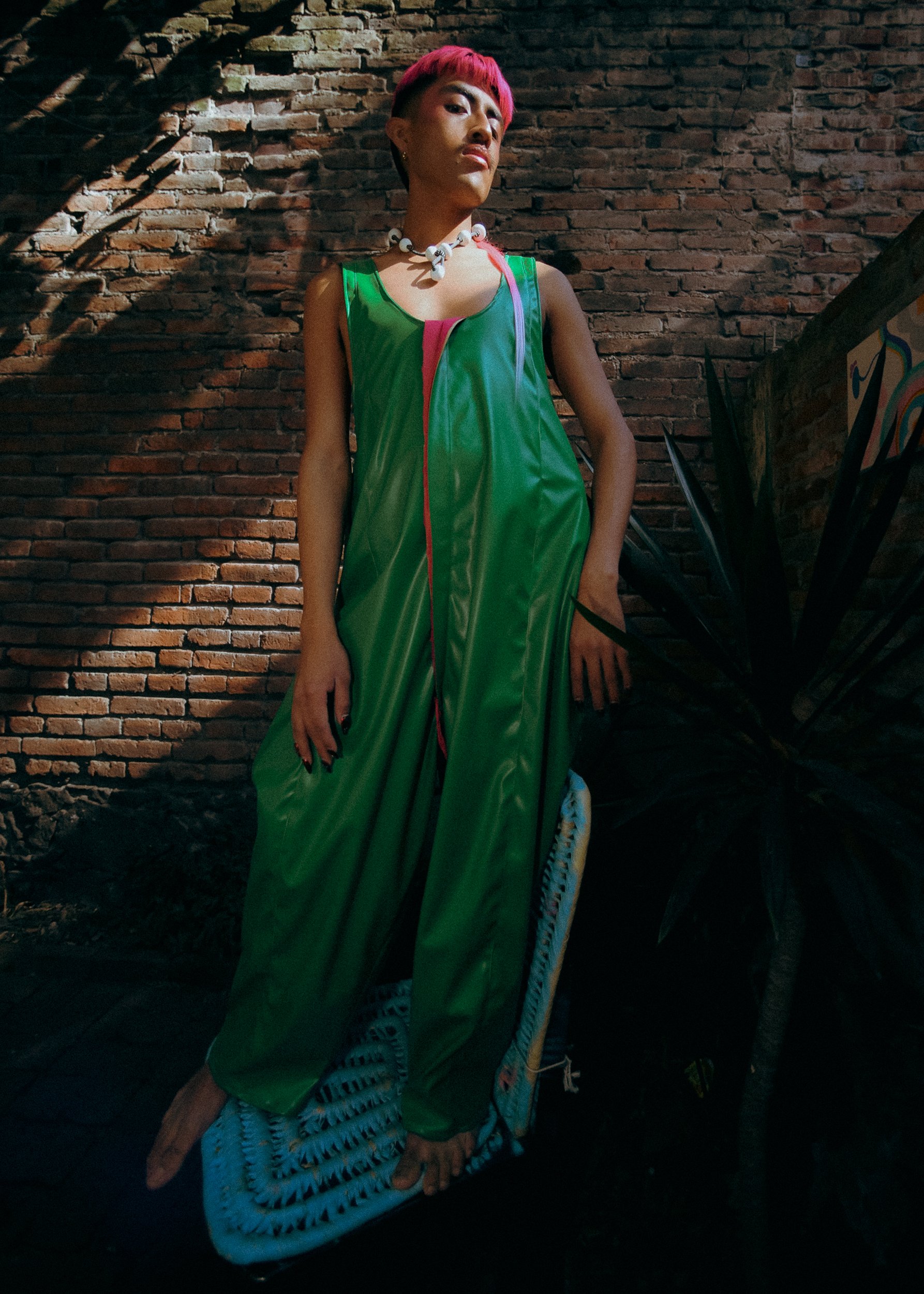 A feminine model with pink hair wearing a green dress and white necklace, standing against a brick wall, with one foot on a chair and shadowed lighting, photographed during a fashion editorial photography session in Los Angeles with fashion photograp