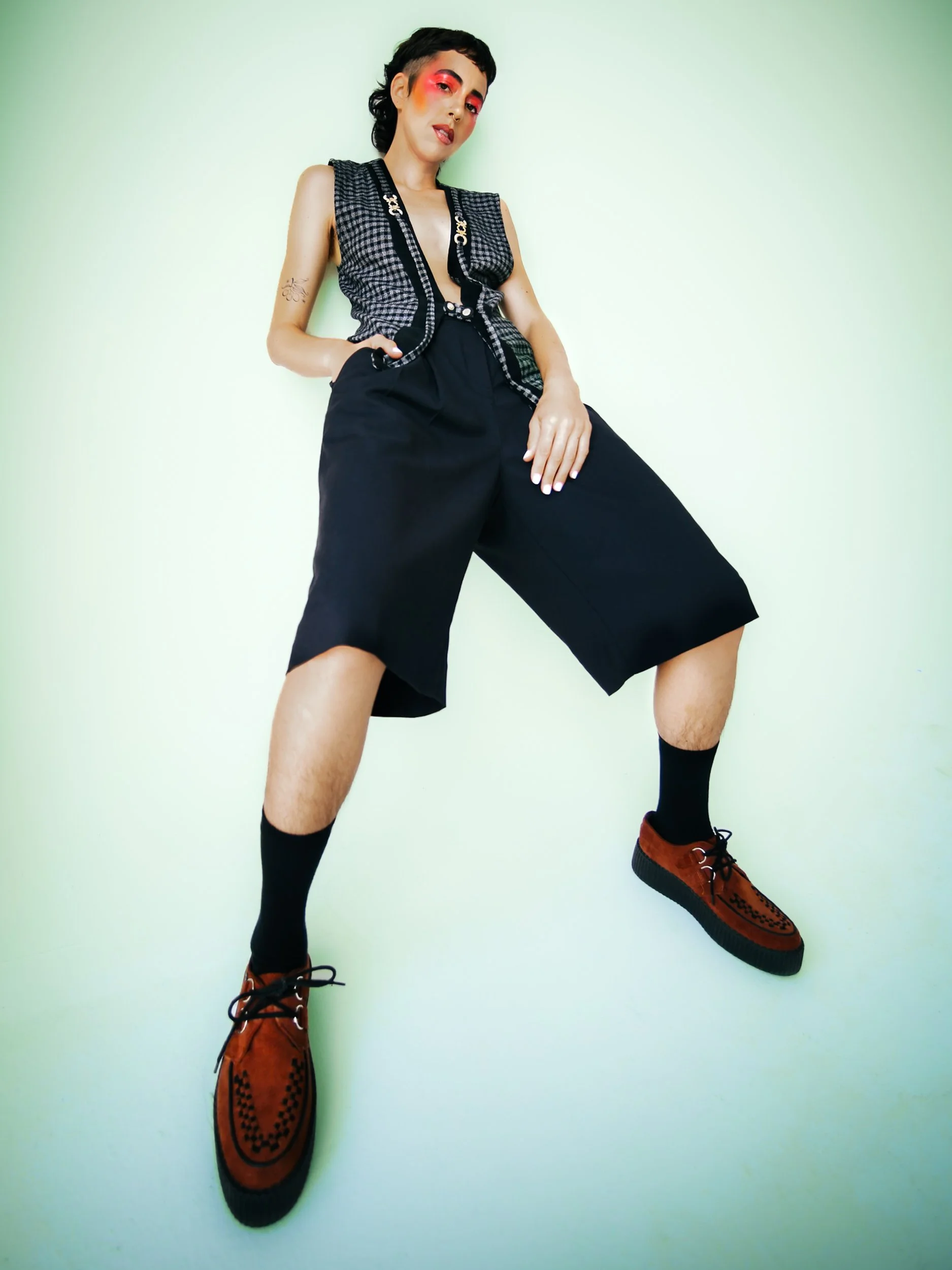 Person crouching on a white floor near a window with light-colored curtains, wearing a checkered sleeveless top and black shorts, with makeup and tattoos., photographed during a studio photography session in Los Angeles for a fashion editorial photos