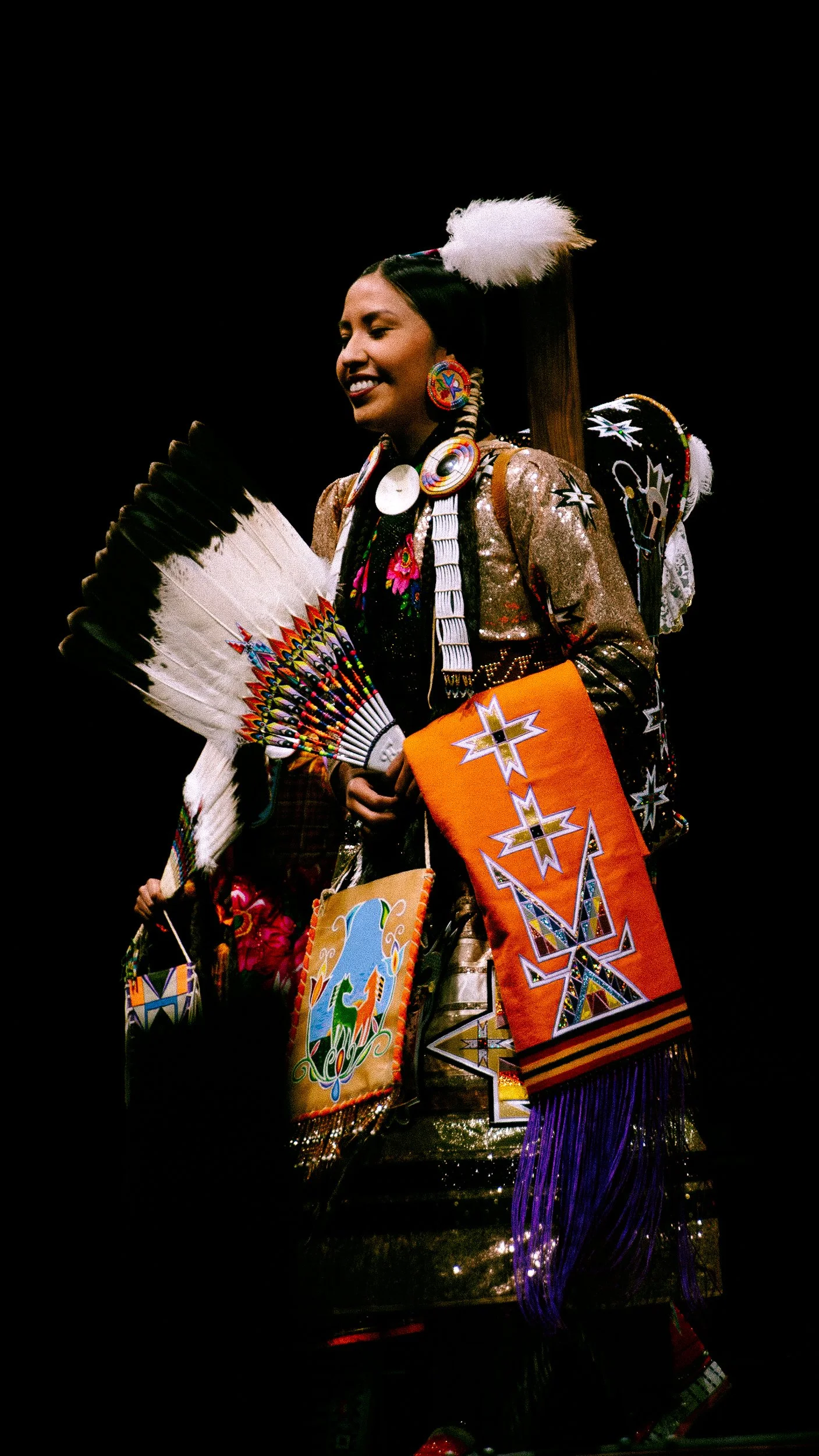 A Native American woman dressed in traditional attire, holding a feather fan, smiling, with intricate beadwork, patterns, and accessories, against a dark background.