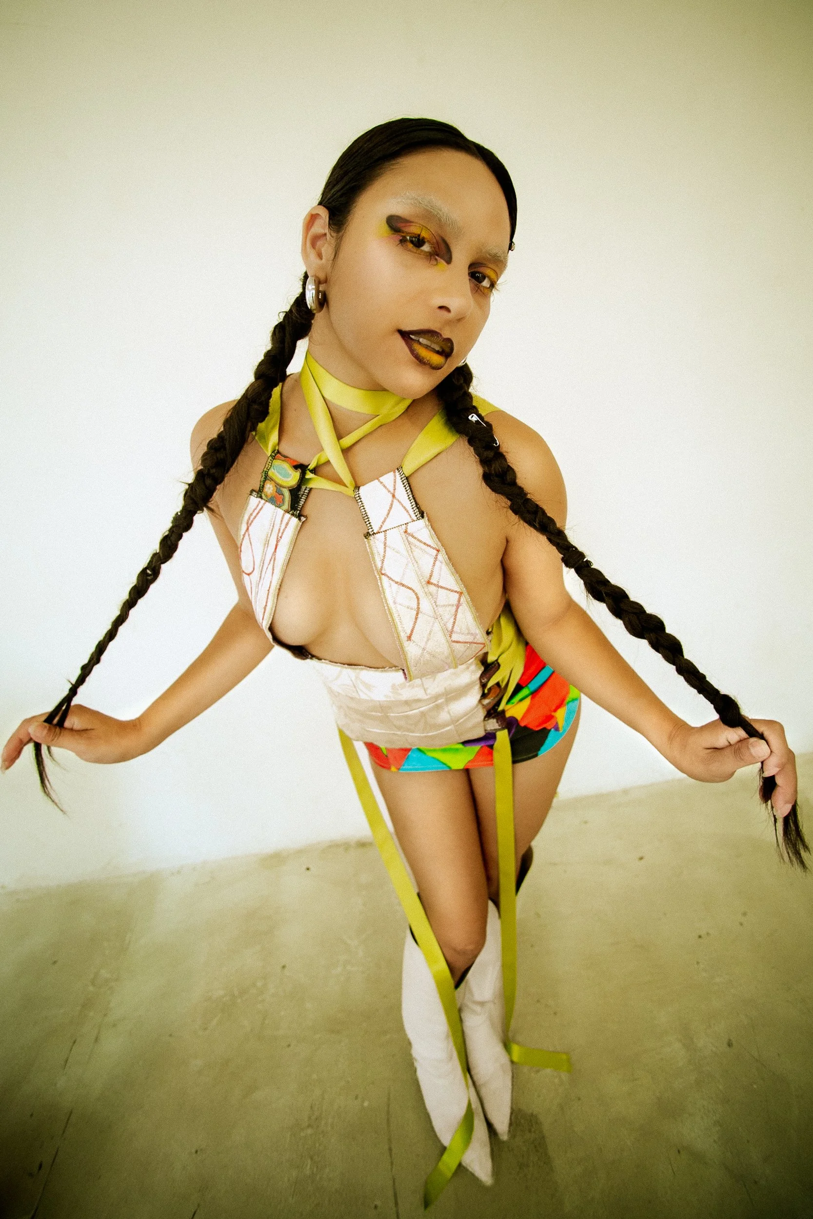 Person with braided hair wearing colorful, bold makeup and a creative, fashion-forward outfit, posing indoors against a plain white wall and glossy concrete floors, shot for an editorial fashion photography session in Los Angeles with fashion photogr