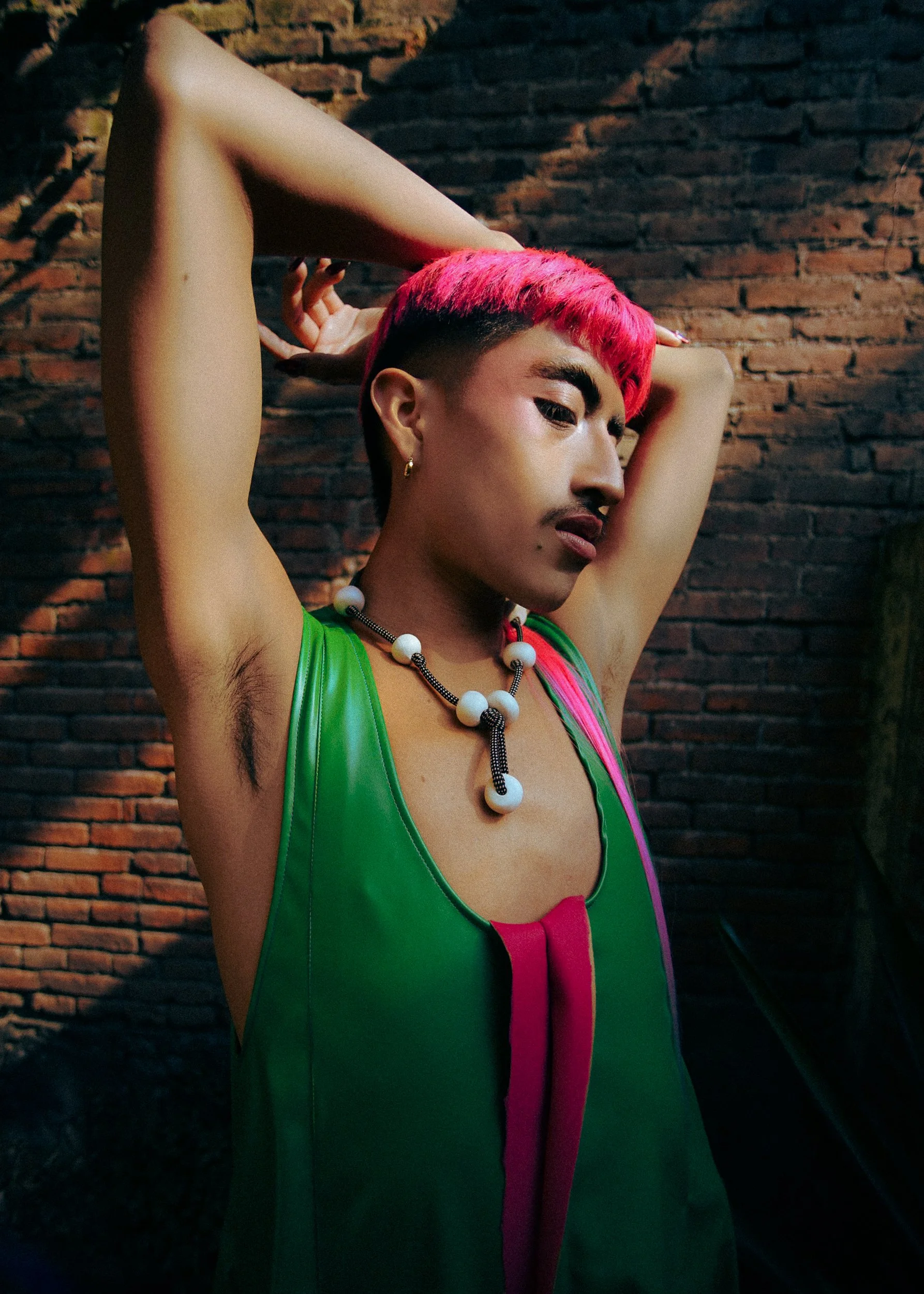 A feminine model  with pink hair styled in a short cut, wearing a green sleeveless top with pink accents and statement jewelry including a beaded necklace. They are posing with arms raised behind the head against a brick wall background, with a serio