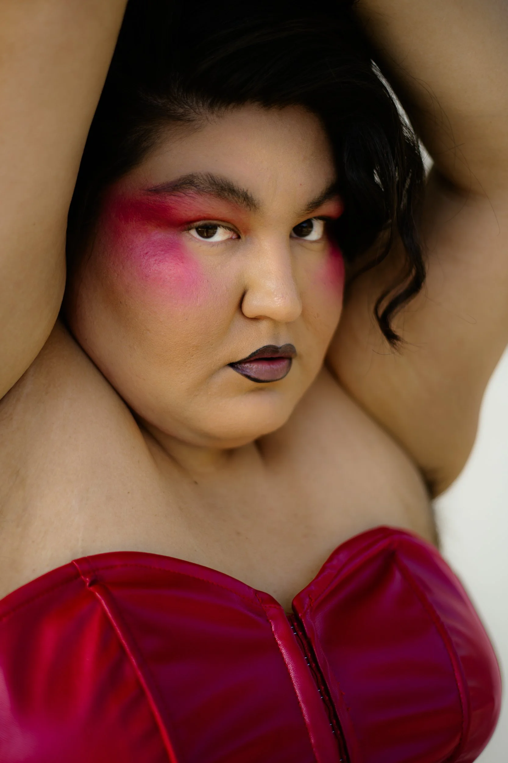 A Latina woman model with bold makeup, including pink blush and eyeshadow, black lipstick, and styled dark hair, wearing a red satin top, photographed during a fashion editorial photography session in Los Angeles with fashion photographer Gí Tenamá..