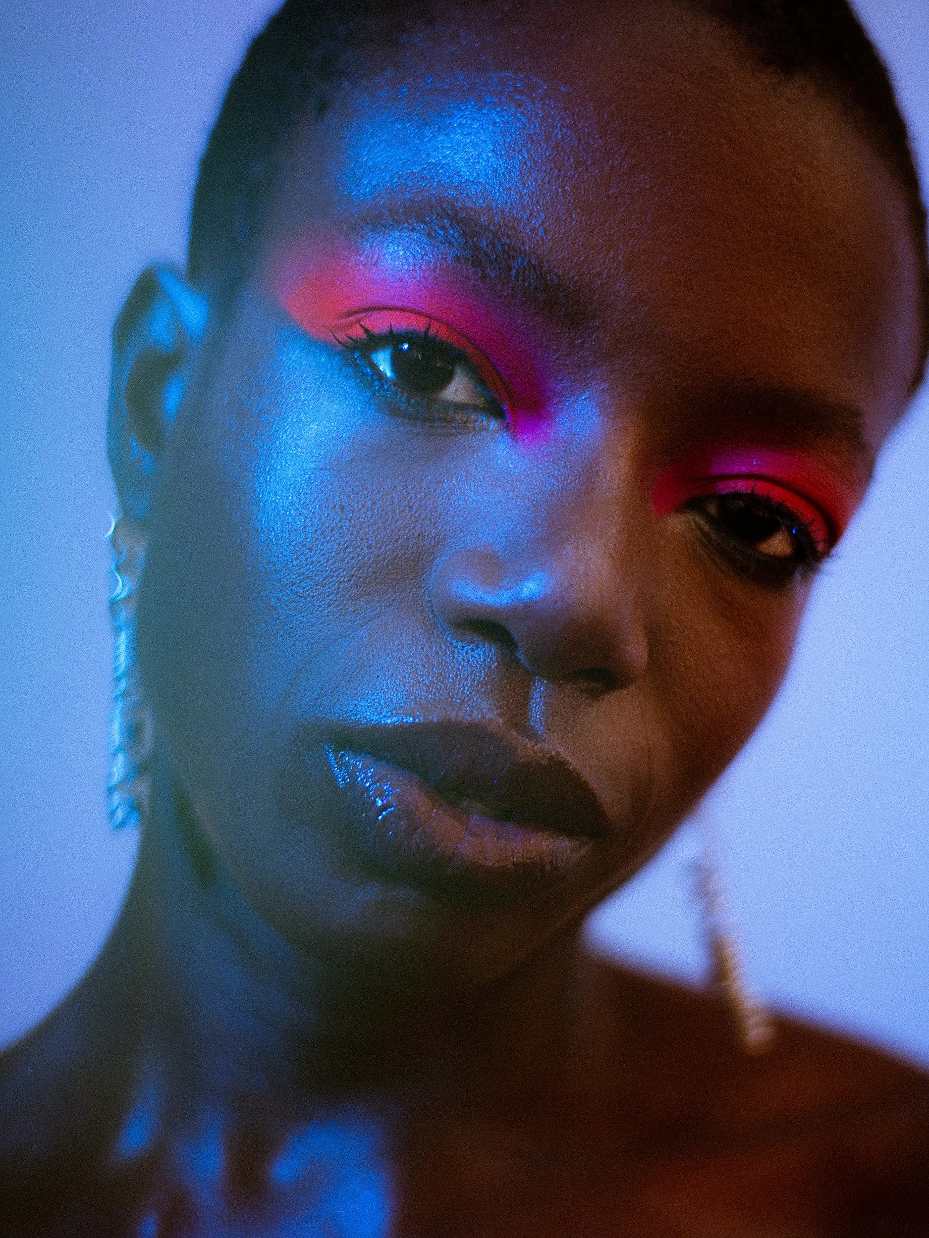 Close-up portrait of a woman model with vibrant pink and blue makeup, wearing earrings, illuminated with colorful lighting, A woman with curly black hair wearing bright makeup, earrings, a rainbow beaded bracelet, and a necklace with a large red hear
