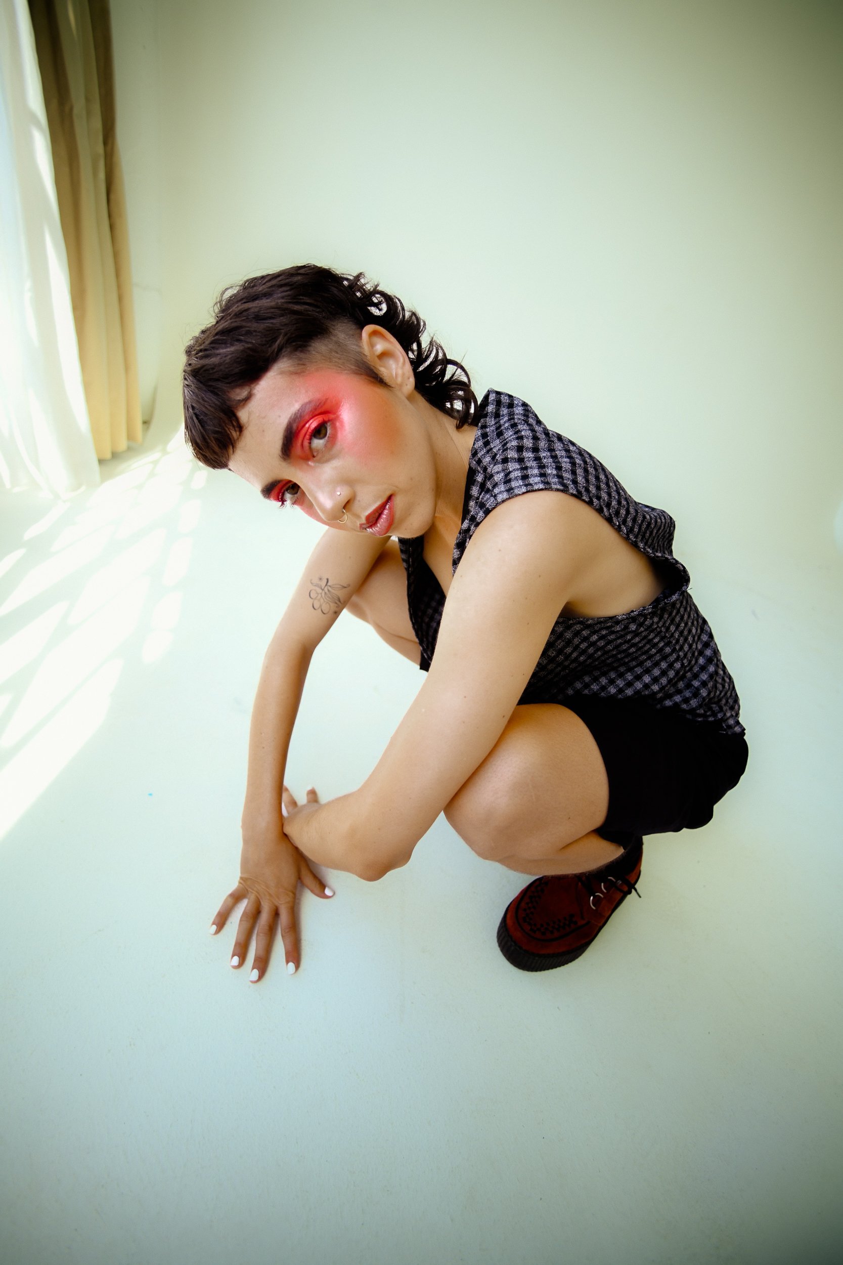 Person crouching on a white floor near a window with light-colored curtains, wearing a checkered sleeveless top and black shorts, with makeup and tattoos., photographed during a studio photography session in Los Angeles for a fashion editorial photos