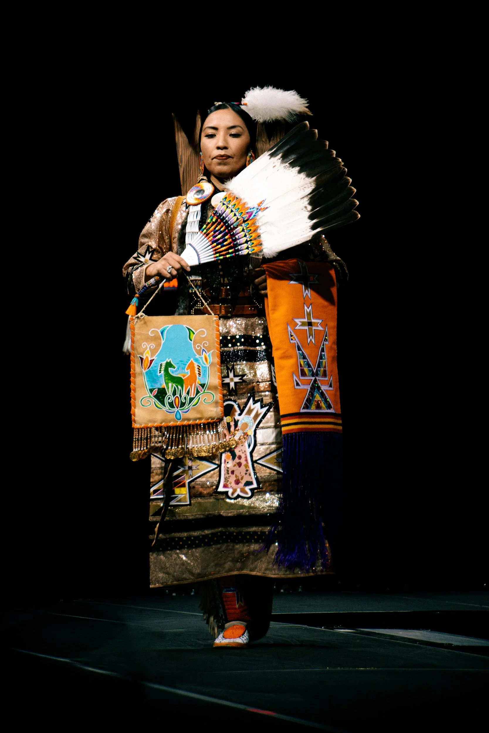 A Native American woman dressed in traditional regalia, holding a feather fan and a decorated bag, standing on a stage with a black background.