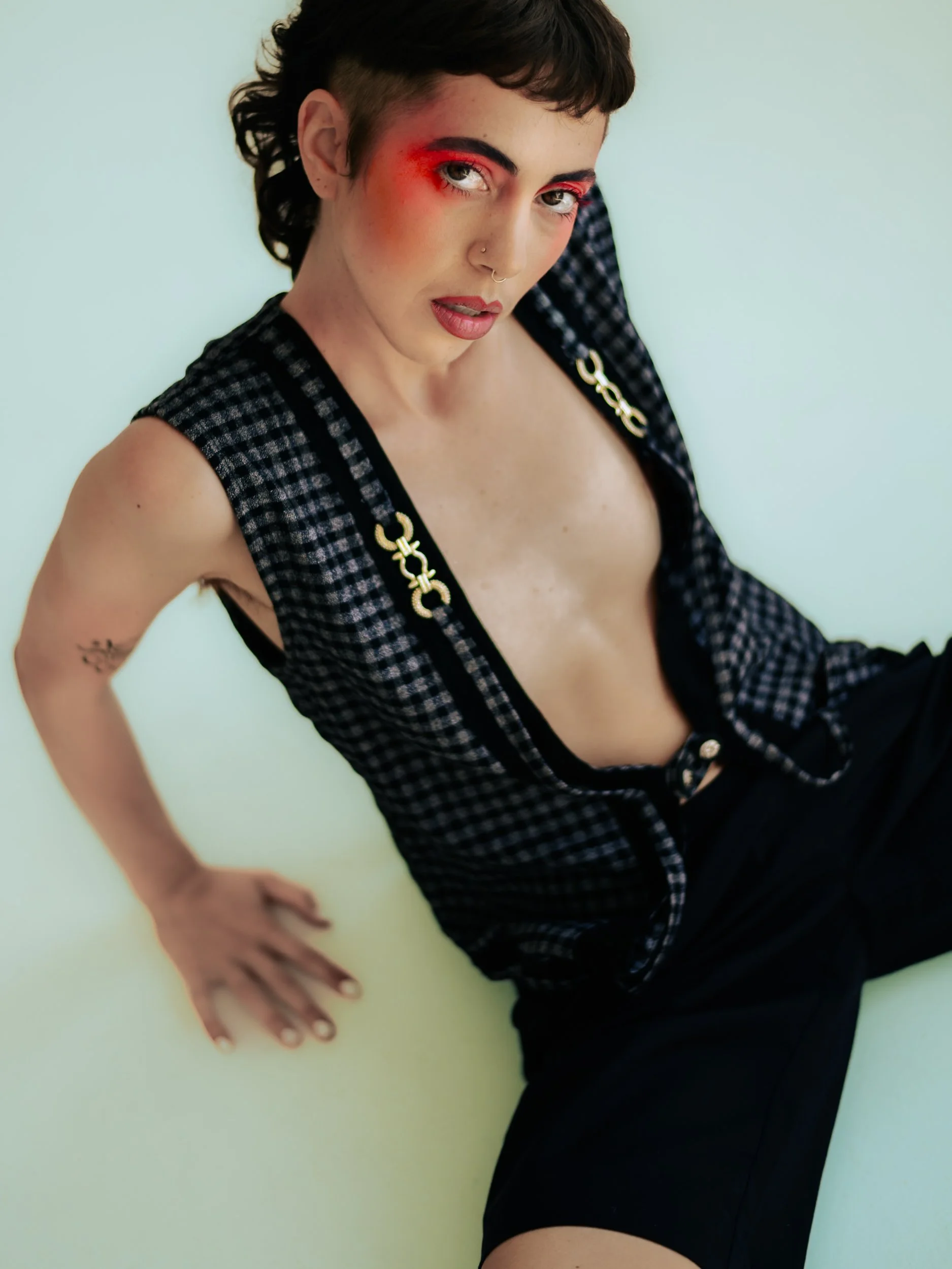 Person crouching on a white floor near a window with light-colored curtains, wearing a checkered sleeveless top and black shorts, with makeup and tattoos., photographed during a studio photography session in Los Angeles for a fashion editorial photos